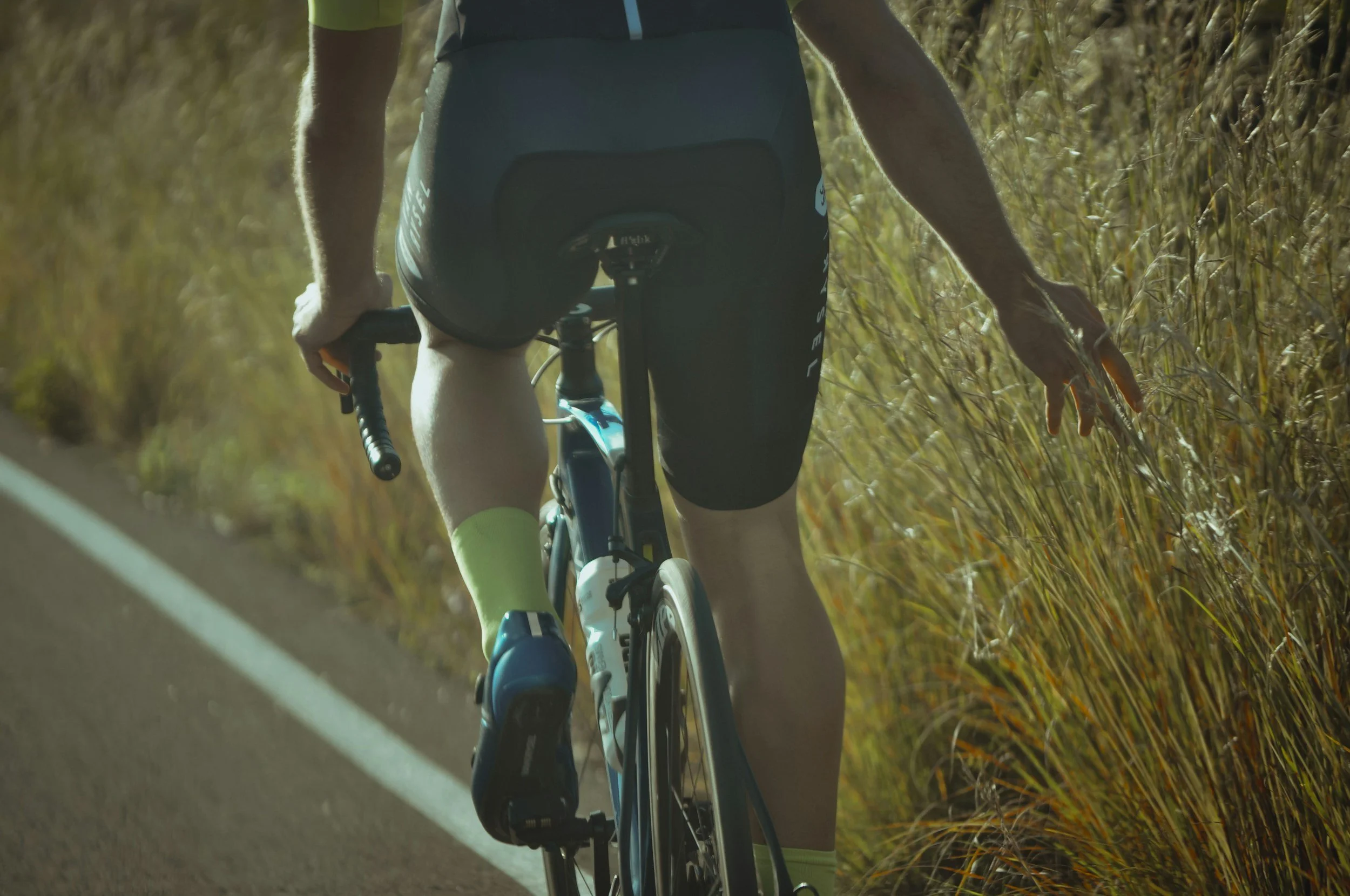 A cyclist riding on a trail through tall grass during daytime, wearing cycling apparel and gloves, viewed from behind.