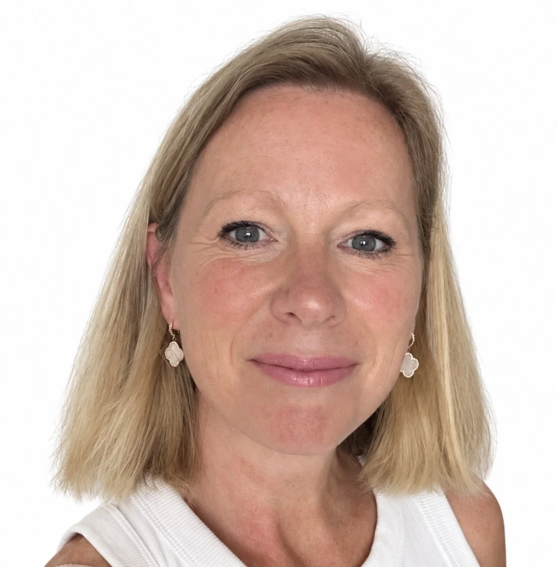 Close-up portrait of a middle-aged woman with shoulder-length blonde hair, wearing earrings and a white sleeveless top, smiling softly against a plain white background.