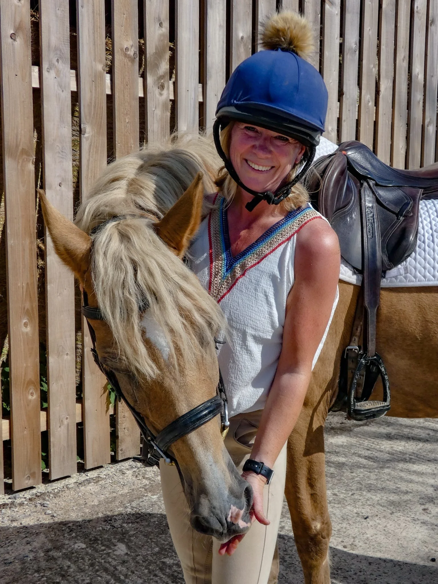 A woman with a big smile wearing a blue riding helmet standing next to a horse, holding its cheek, in an outdoor area with a wooden fence in the background.