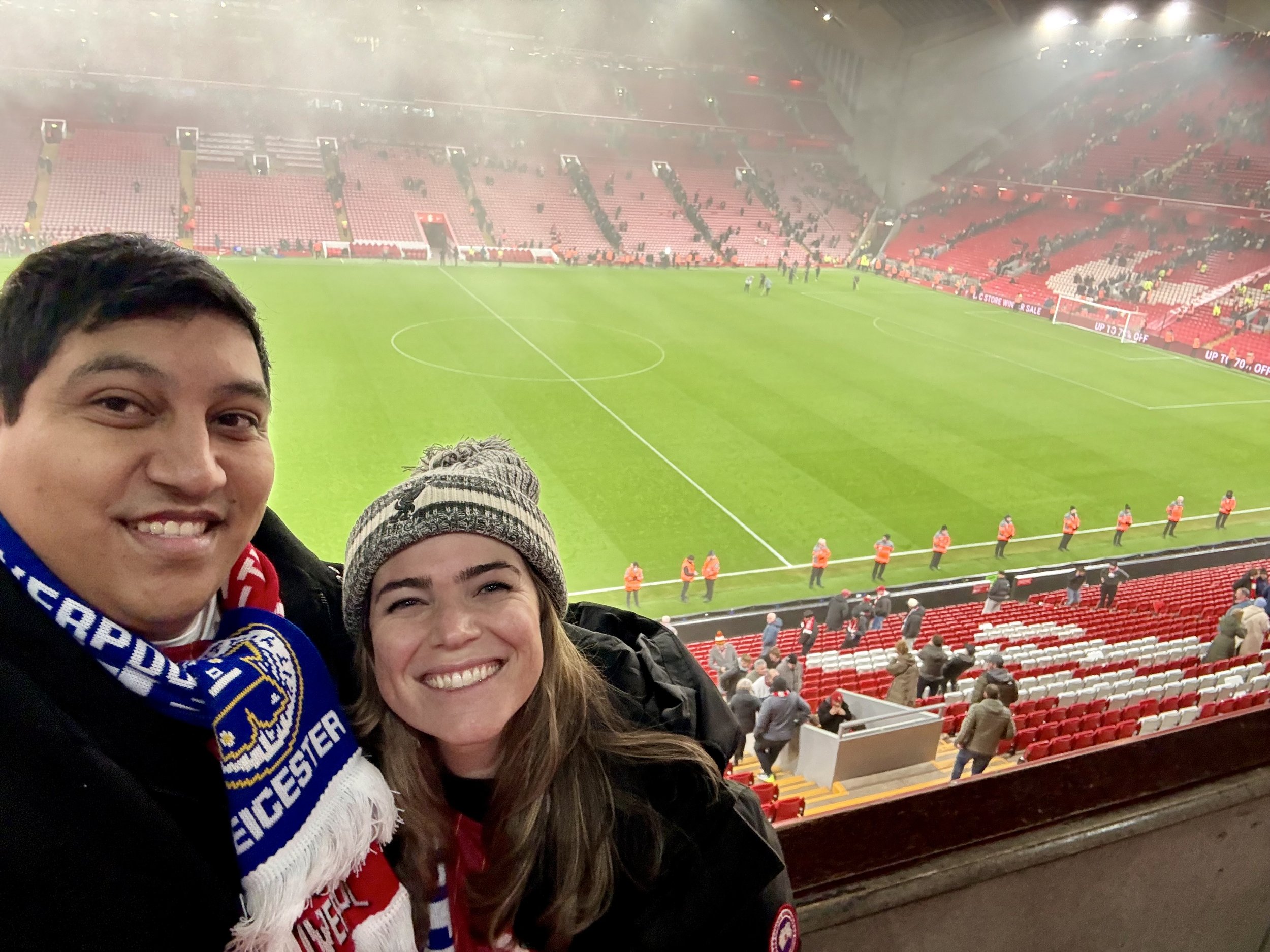 Two smiling women at a soccer stadium with a mostly empty red seating area and some spectators. The field is visible in the background.