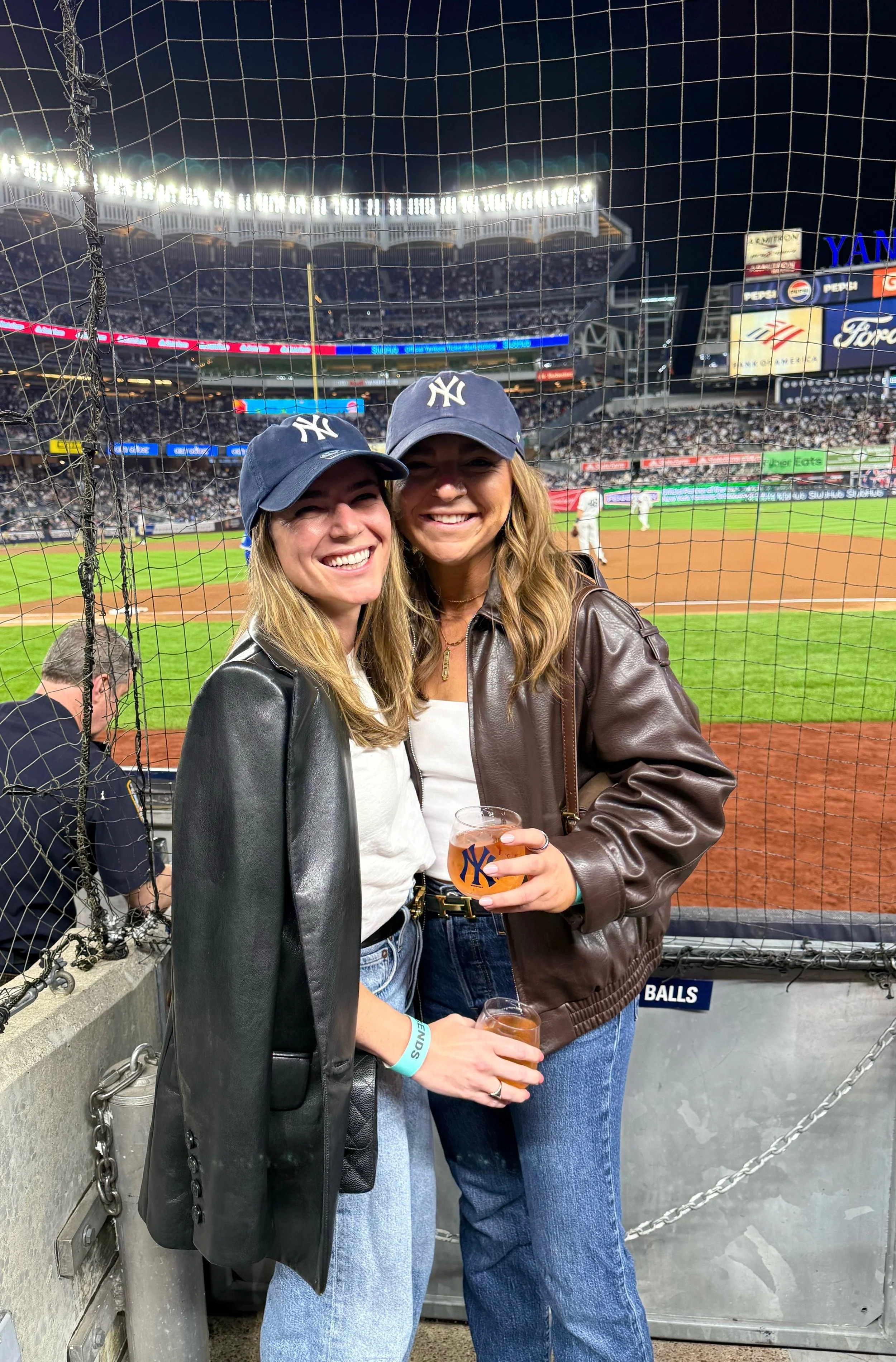 Two women wearing New York Yankees hats smiling at a baseball game, holding drinks, standing behind a net at a stadium.