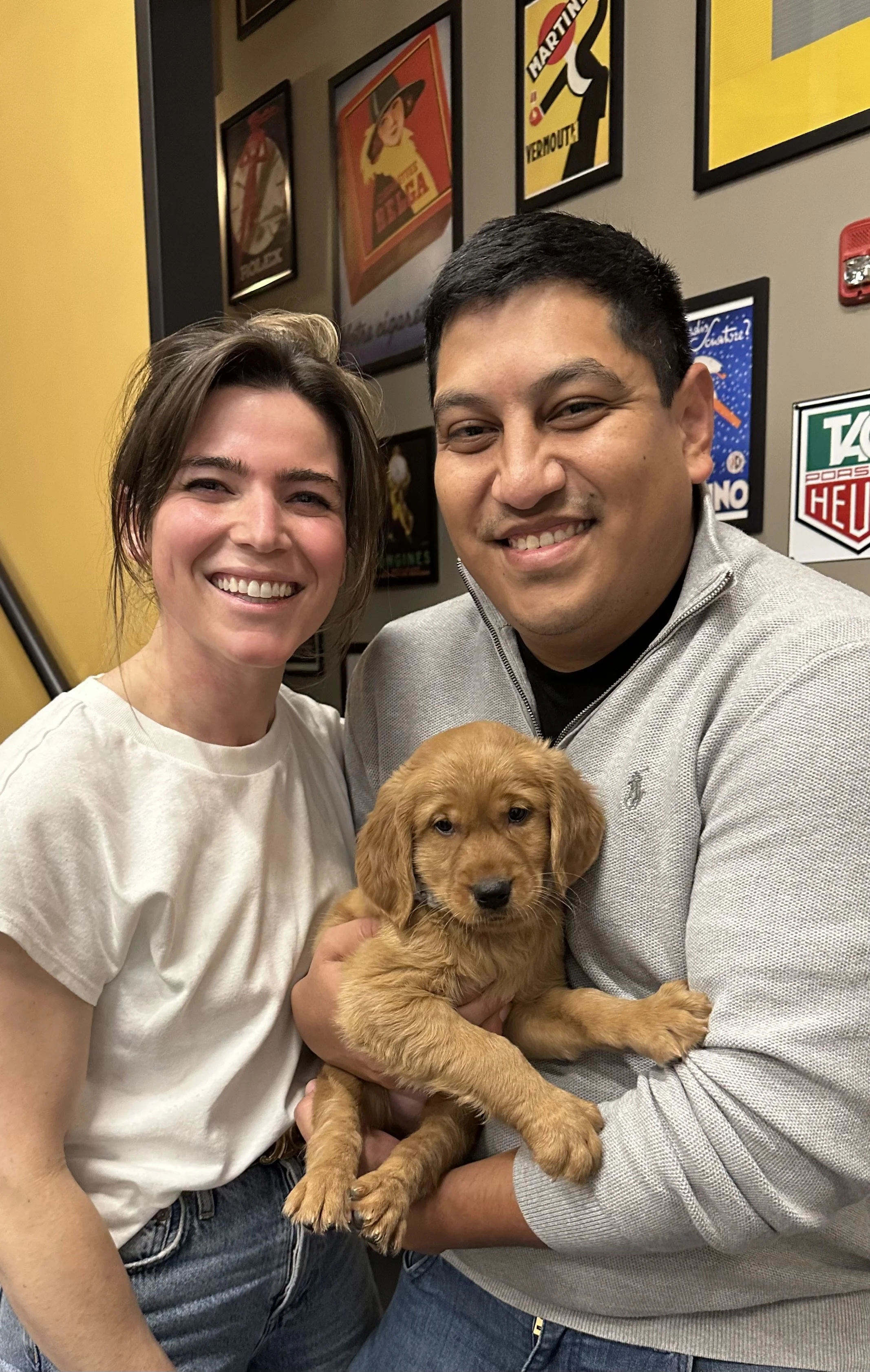 Two smiling people, a woman and a man, holding a golden retriever puppy inside a room with plaques and posters on the wall.