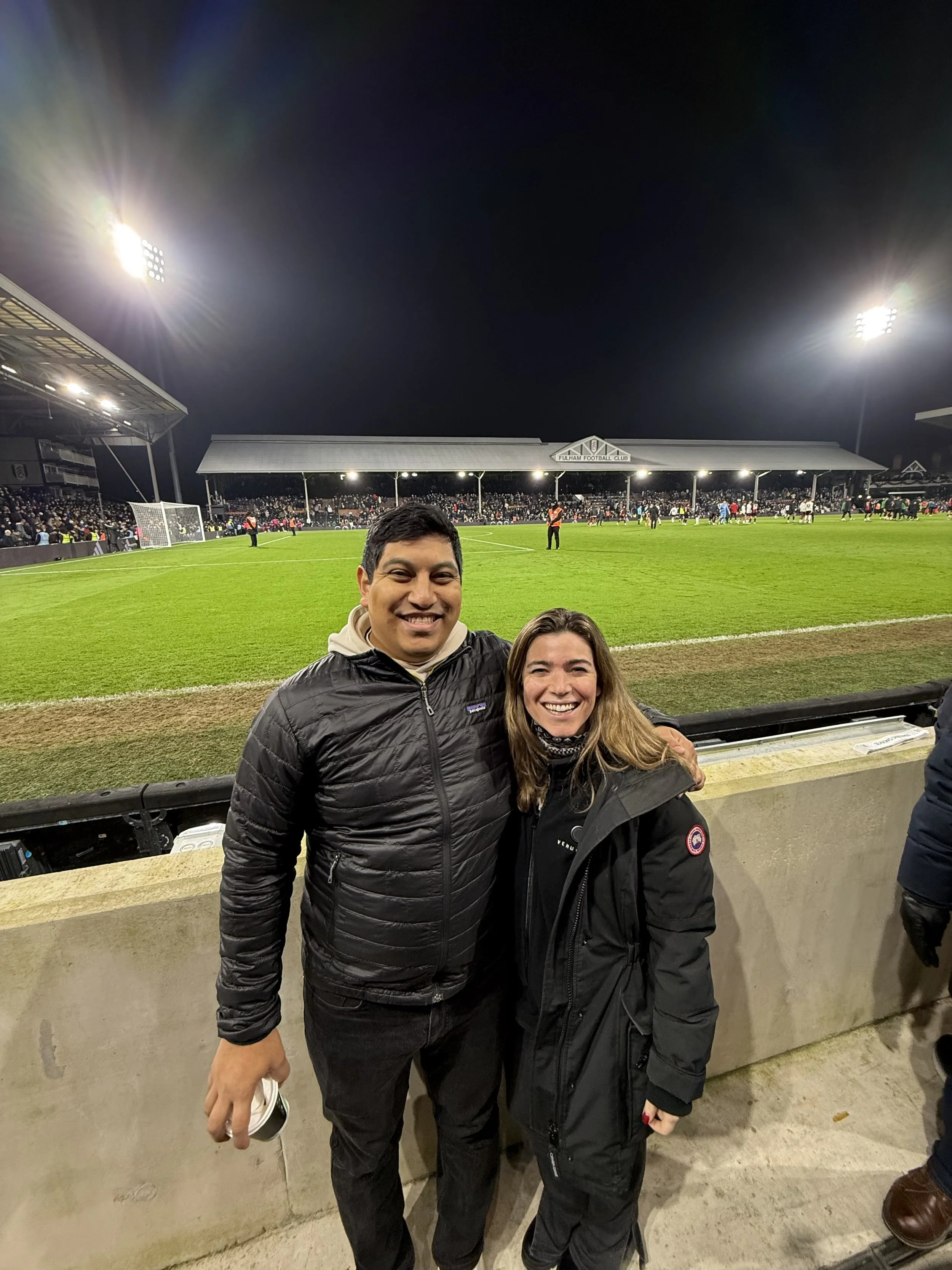 Two smiling people, a man and a woman, standing at a night football match at Fulham Football Club stadium. The stadium is lit up and filled with spectators.