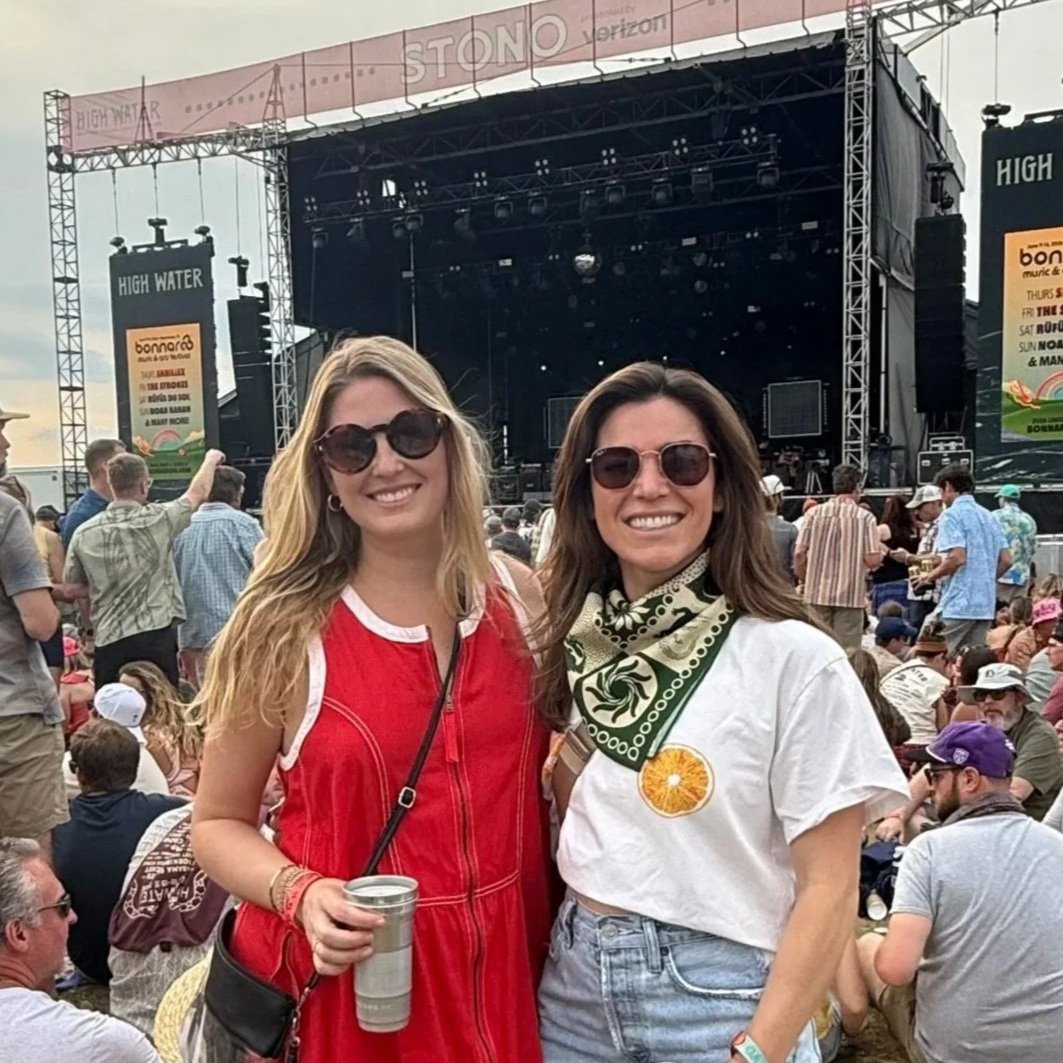 Two women wearing sunglasses at a music festival with a stage in the background, surrounded by a crowd of people.