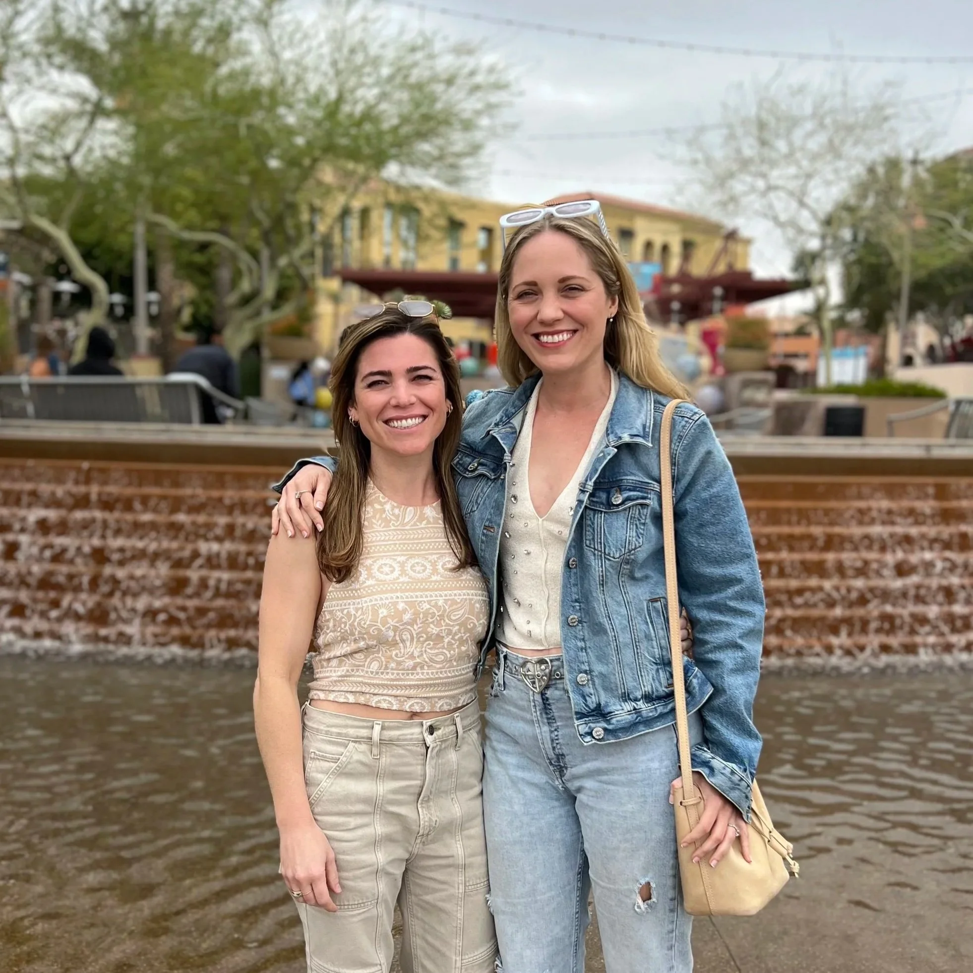 Two smiling young women standing outdoors by railroad tracks with trees and buildings in the background. They are wearing casual clothes, sunglasses on their heads, and are close together with their arms around each other.