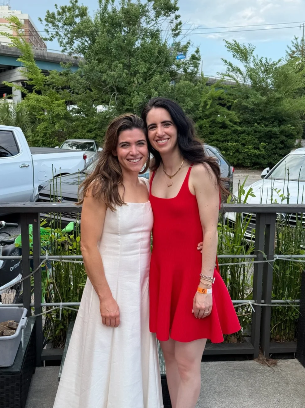 Two women standing outdoors, smiling, with trees, parked cars, and a highway in the background, posing for a photo together.