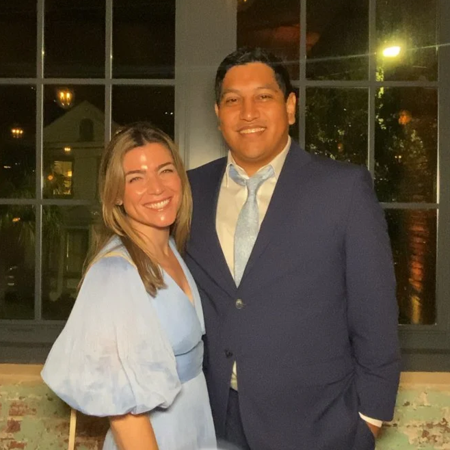 A woman and man smiling and posing together indoors near a window at night, dressed in formal attire.