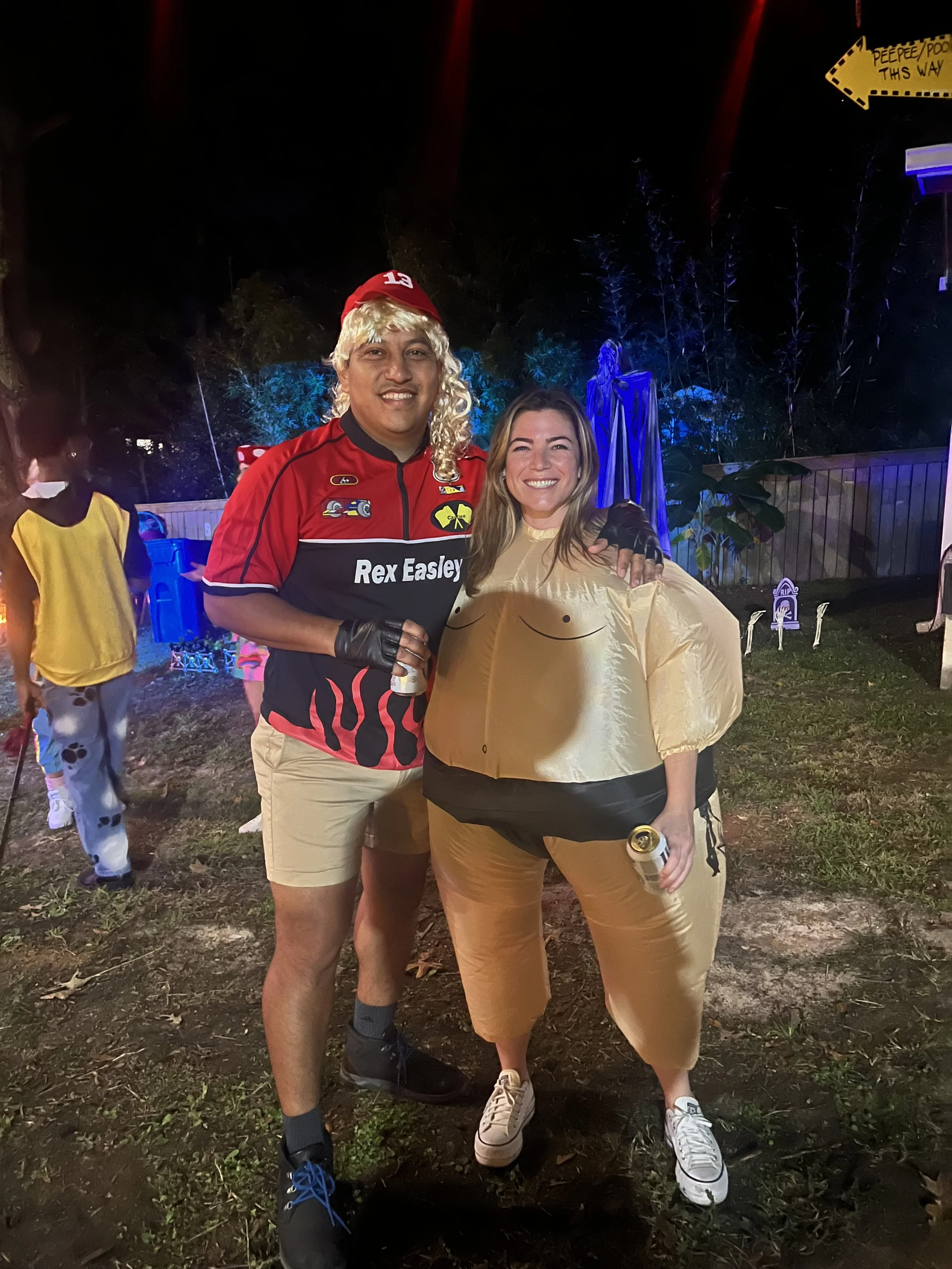 Two people smiling, standing outdoors at night with colorful lights and a small garden behind them. One person is dressed in costume with a blond wig and red hat, the other is wearing yellow and holding a drink.