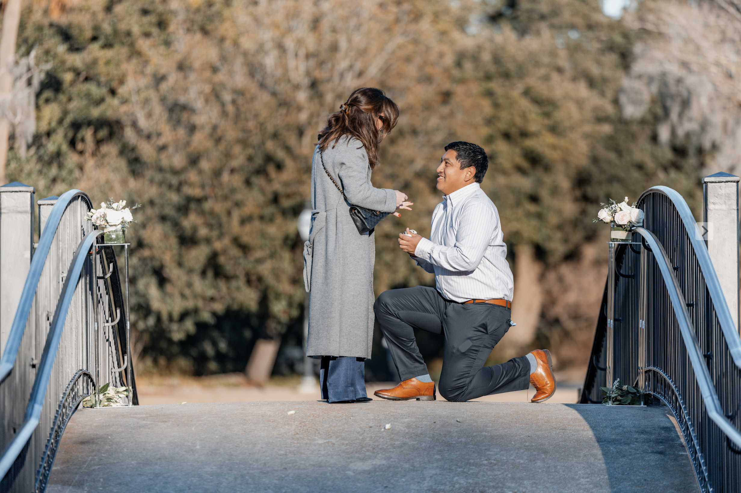 A man kneeling on one knee proposing to a woman on a bridge decorated with flowers during autumn, with trees in the background.