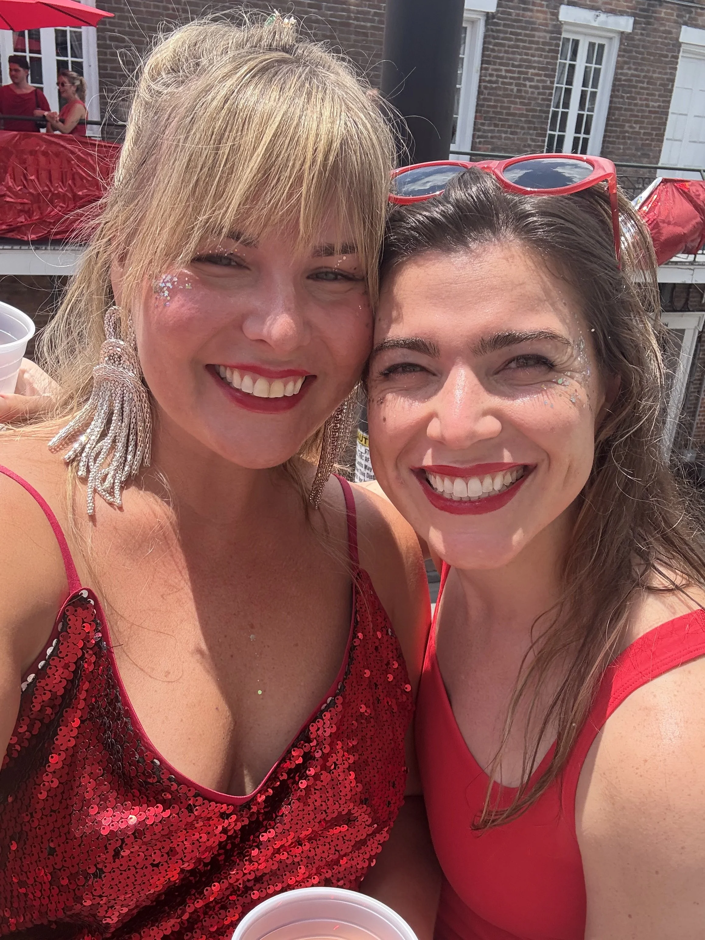 Two women dressed in red, smiling and taking a selfie, with a brick building and people in the background at an outdoor event.
