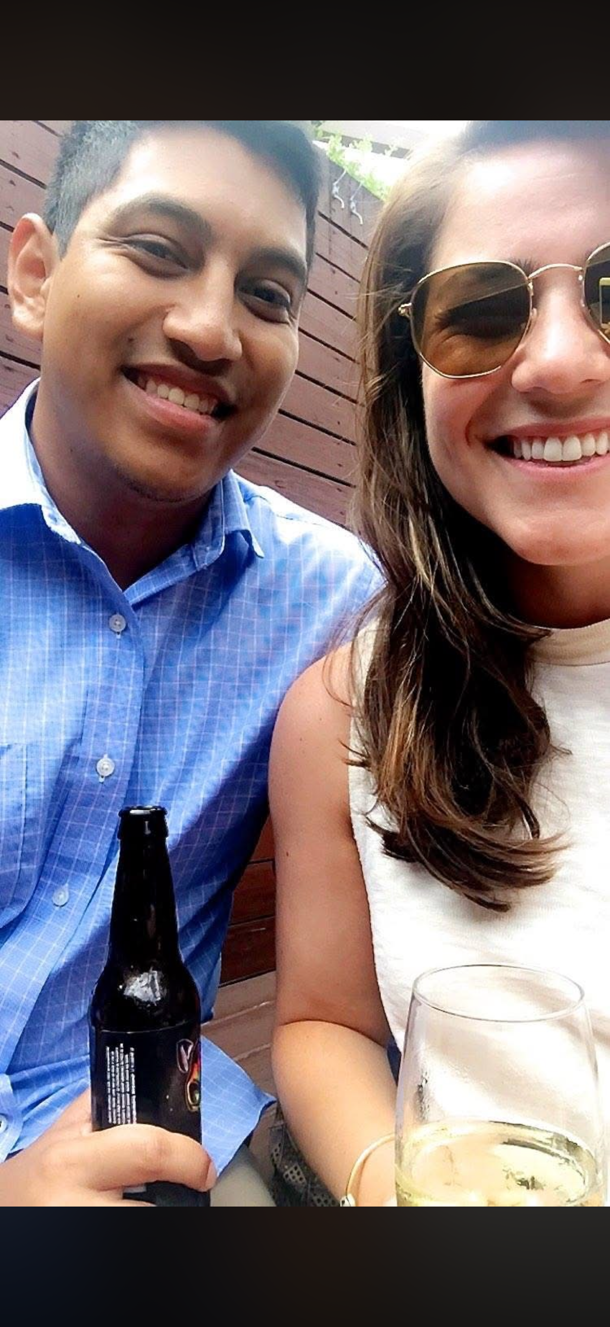 A smiling man in a blue checkered shirt holding a beer bottle and a woman wearing sunglasses and a white sleeveless top enjoying drinks in an outdoor setting with a wooden fence in the background.