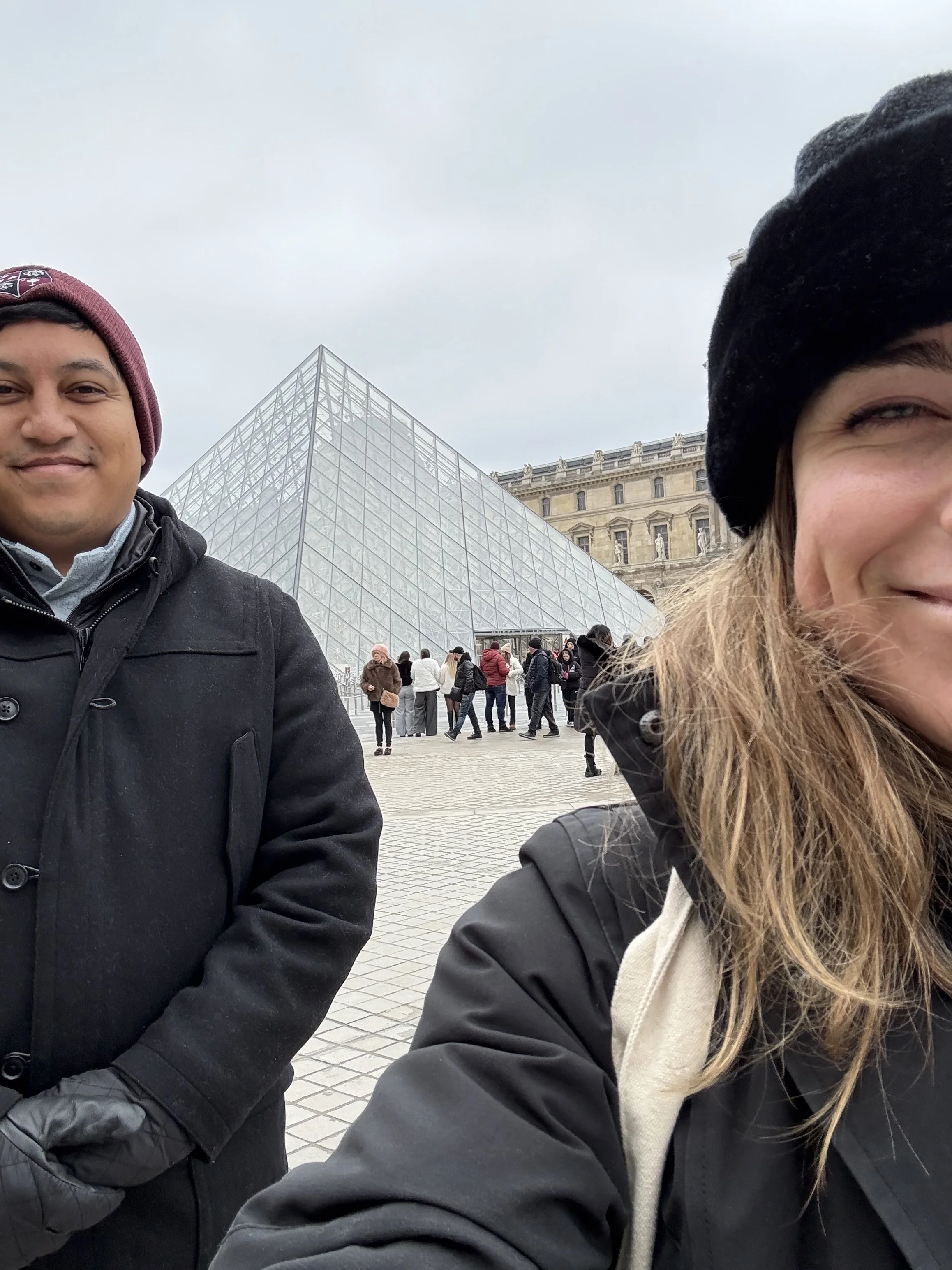 Two people taking a selfie in front of the Louvre Museum in Paris, France; one man smiling on the left side and a woman with blonde hair on the right side, both wearing warm clothing and beanies.