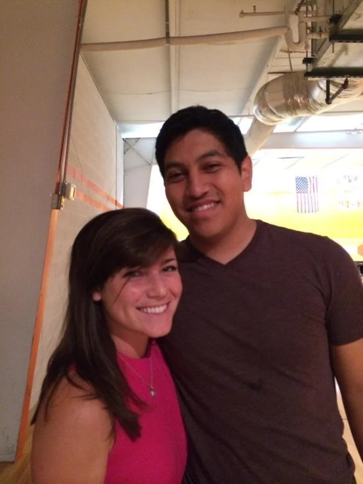 A young woman in a pink top smiling and a young man in a dark shirt smiling, standing close together indoors with an American flag in the background.