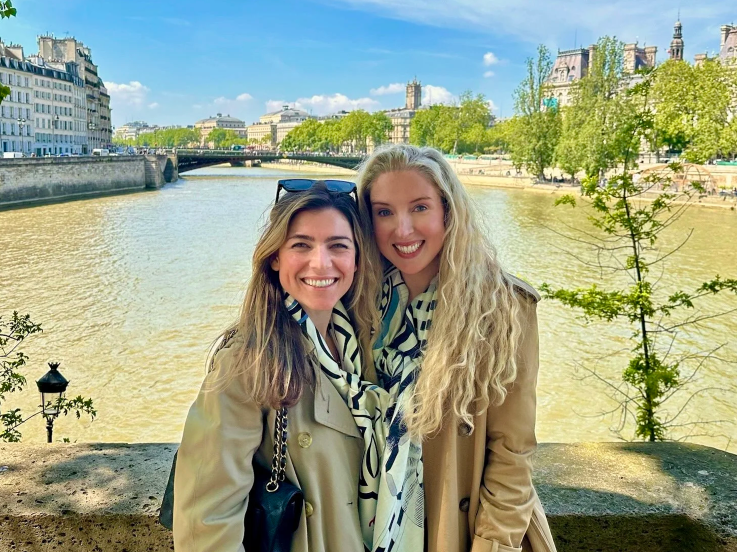 Two smiling women standing outdoors by a river, with city buildings and green trees in the background on a sunny day.
