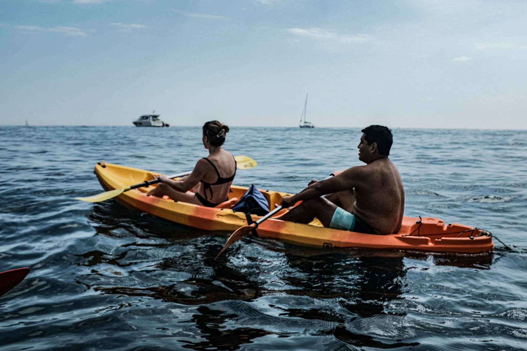 A woman and man on a kayak on the ocean, with boats and a sailboat in the background, under a partly cloudy sky.