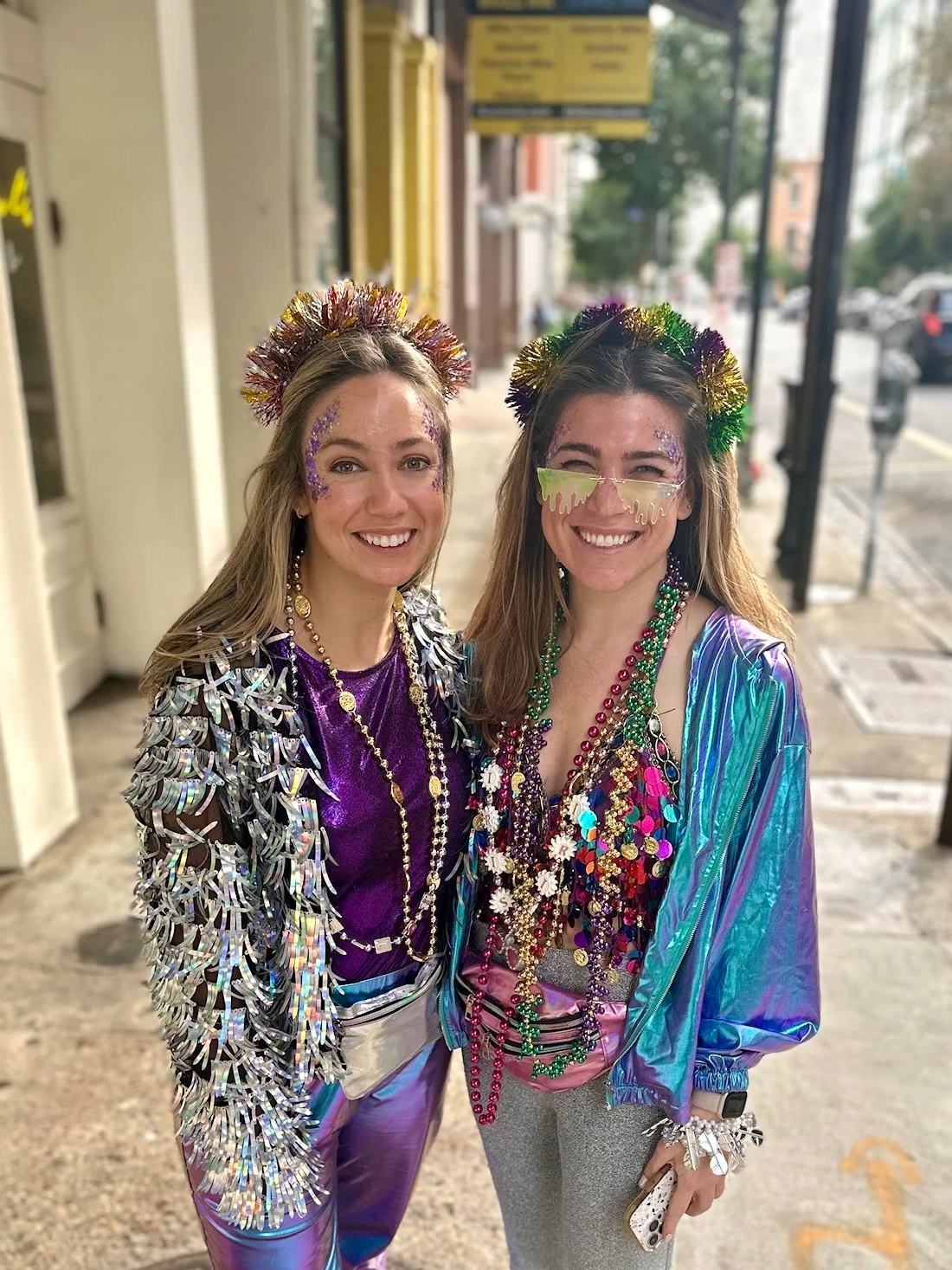 Two women wearing colorful, shiny costumes with beads and necklaces, standing on a sidewalk during the day, celebrating Mardi Gras with festive accessories and glitter face paint.