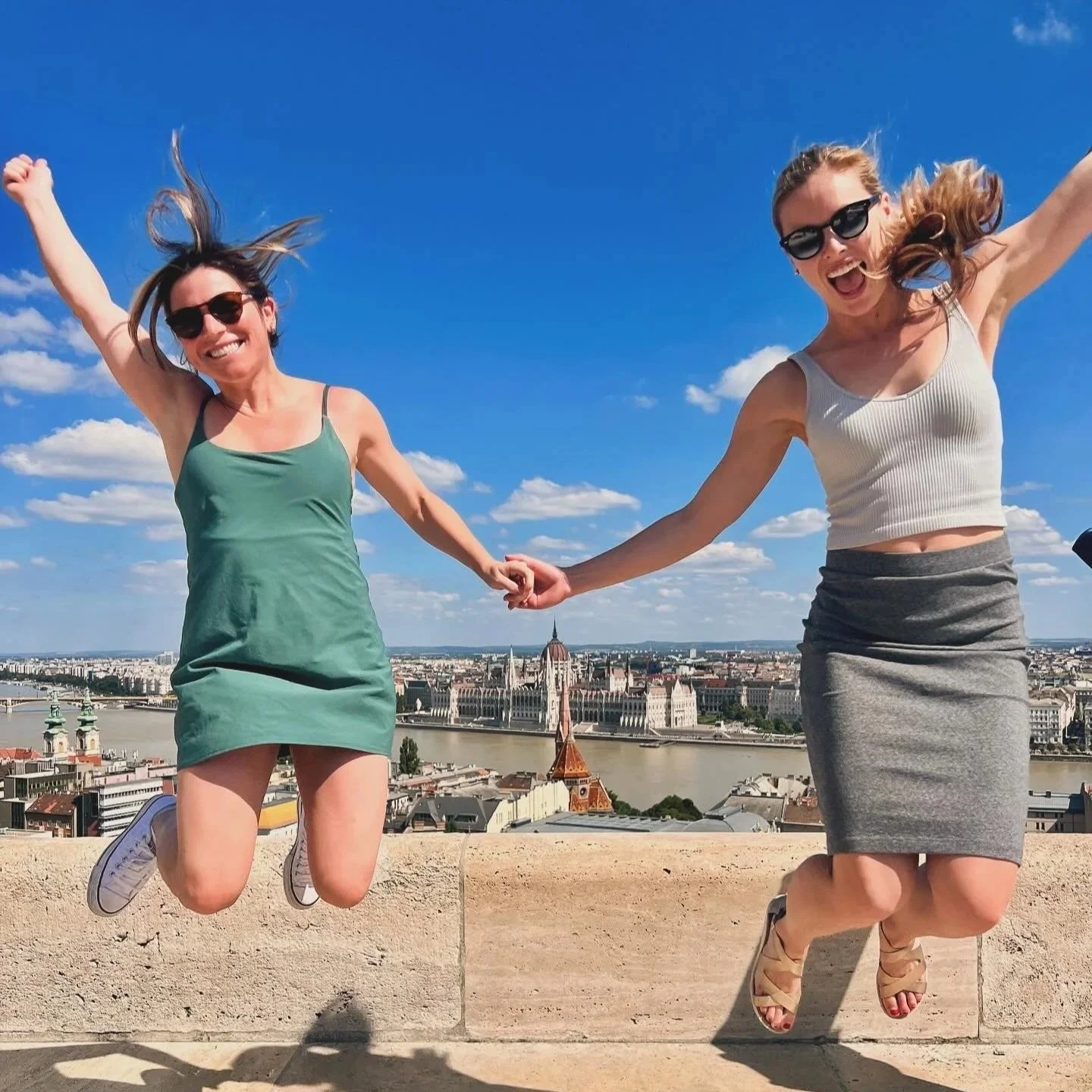 Two women jumping in the air while holding hands with a cityscape and river in the background, under a blue sky with clouds.