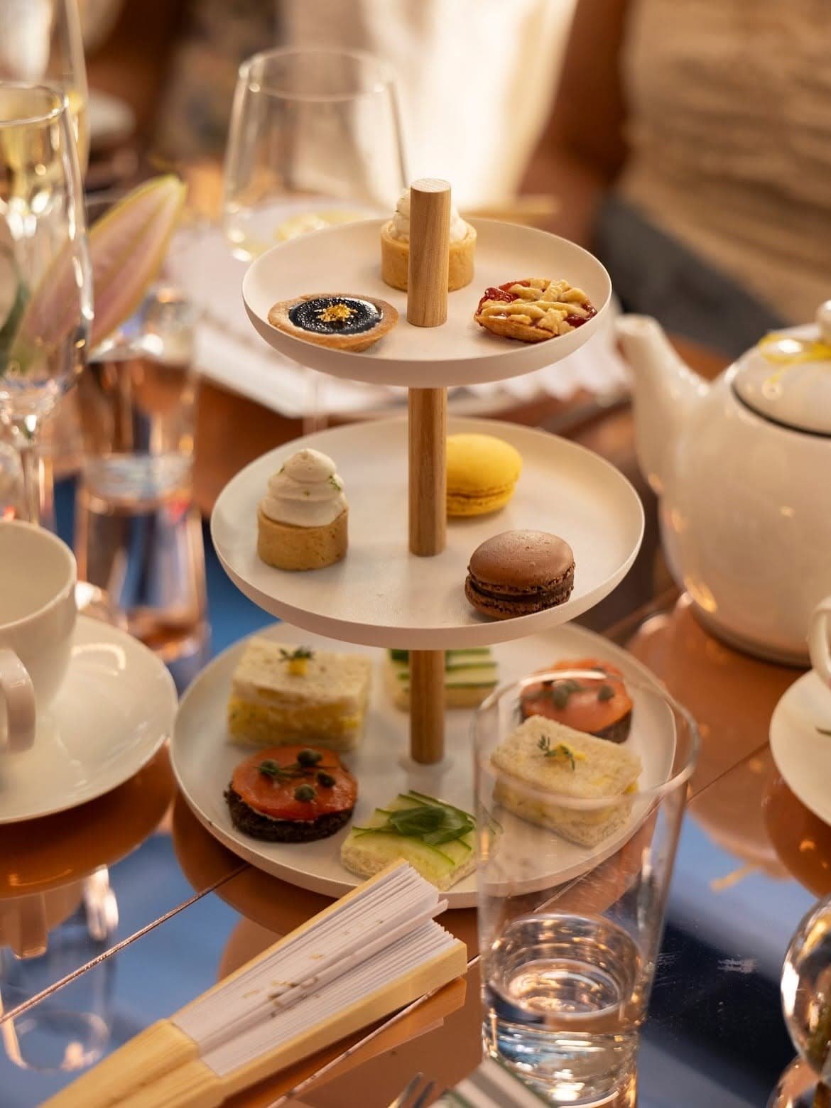 Multi-tiered serving tray high tea at Carton House NYC with assorted small desserts and finger foods, surrounded by teacups, glasses, and utensils on a dining table.