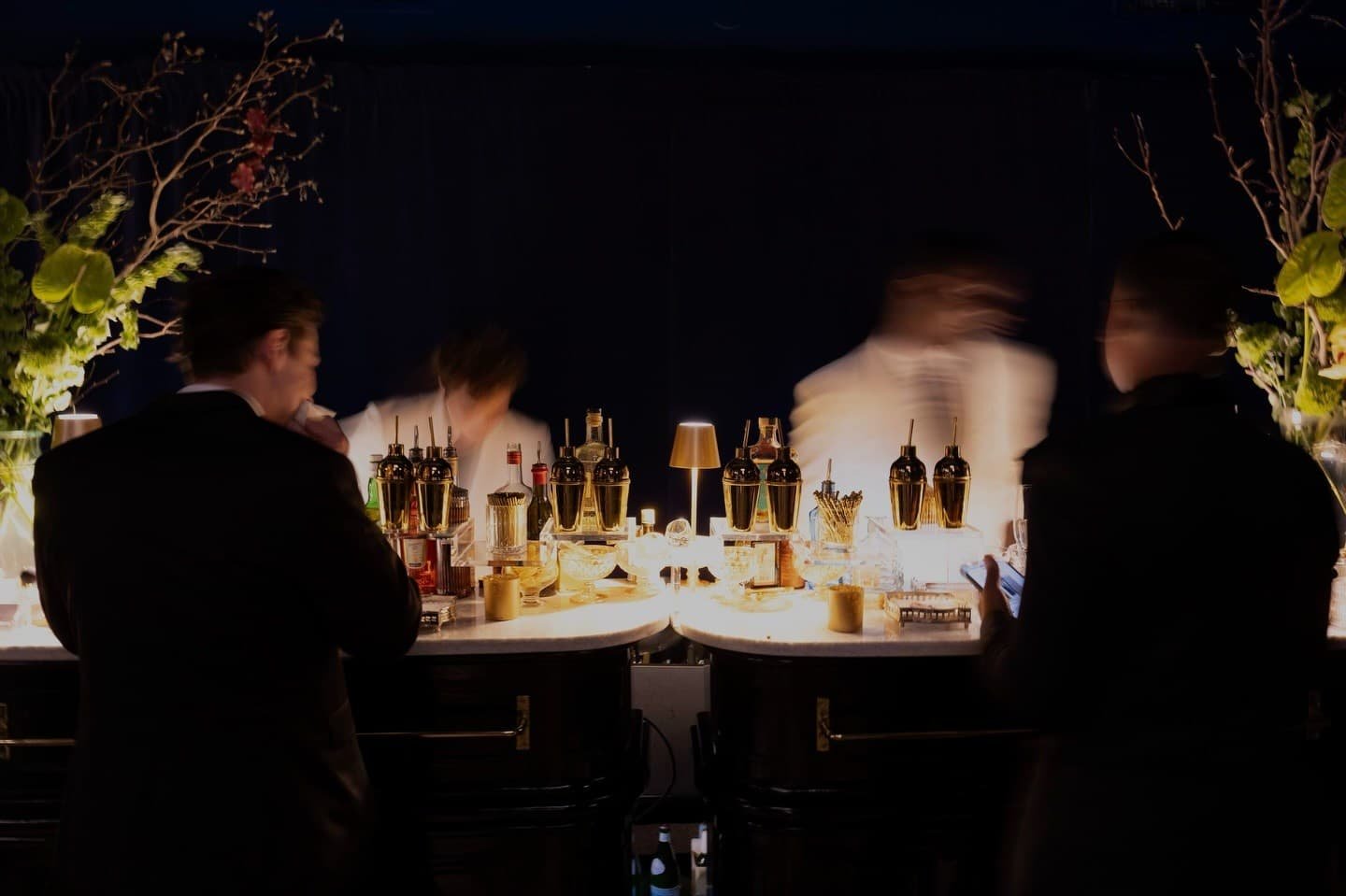 People socializing at a bar, with bottles and mixers on the counter, dim lighting, and floral decorations on the sides.