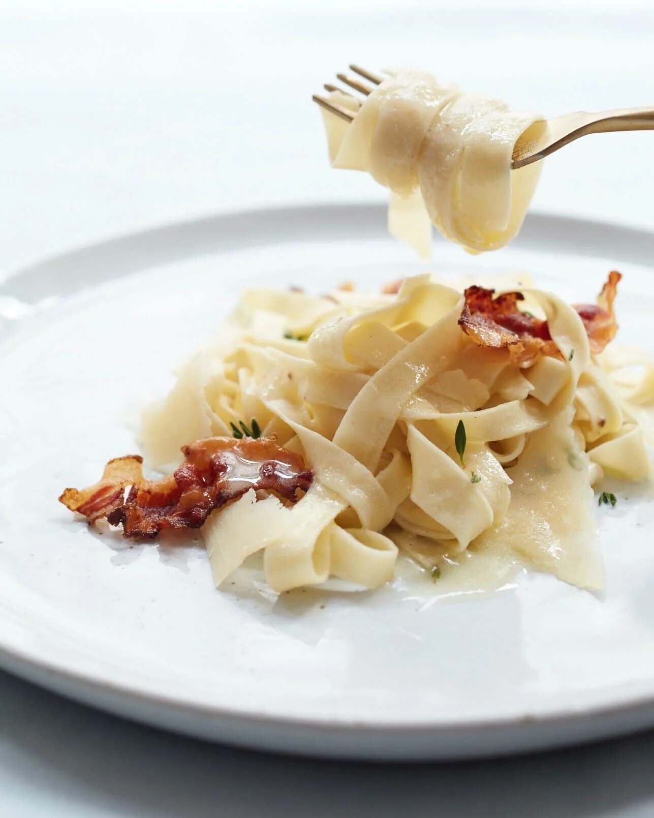 Plate of fettuccine pasta with bacon and herbs, with a fork lifting a piece of pasta.
