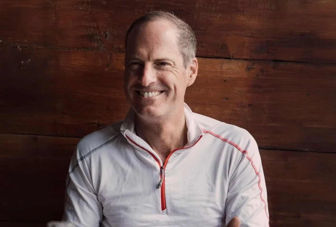 Smiling guest in a white jacket seated against a wood-paneled wall.
