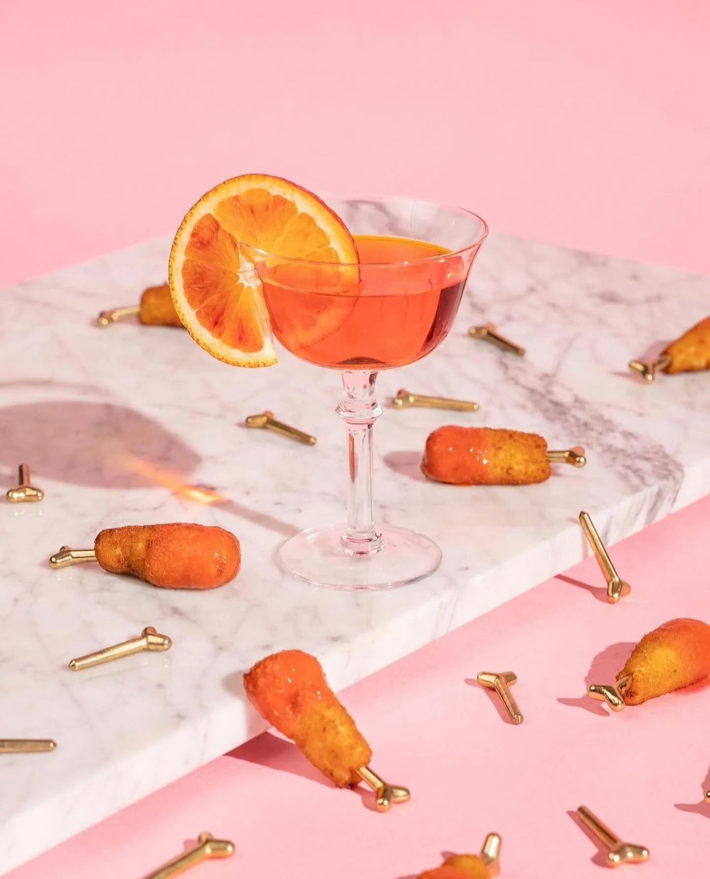 Pink cocktail with orange garnish surrounded by miniature carrot-shaped hors d’oeuvres.