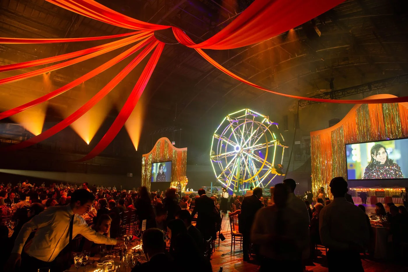 Large-scale indoor celebration with a Ferris wheel installation, red draping, screens, and guests.