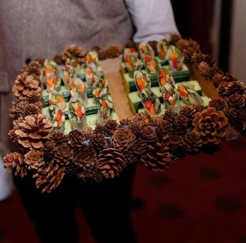 Sushi rolls topped with vegetables on a rustic board edged with pinecones.