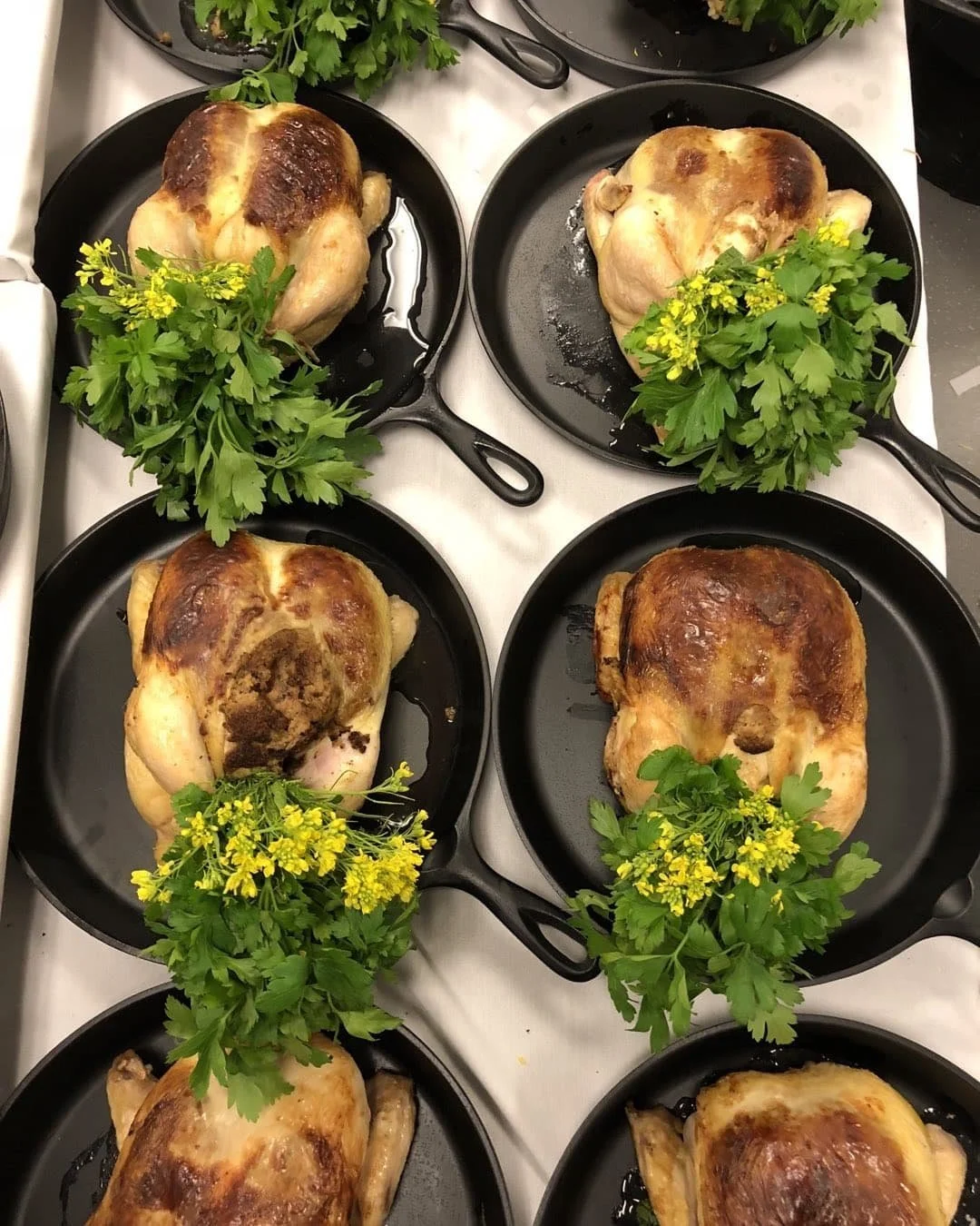 Roasted chickens presented in individual cast-iron skillets with herbs and flowers.