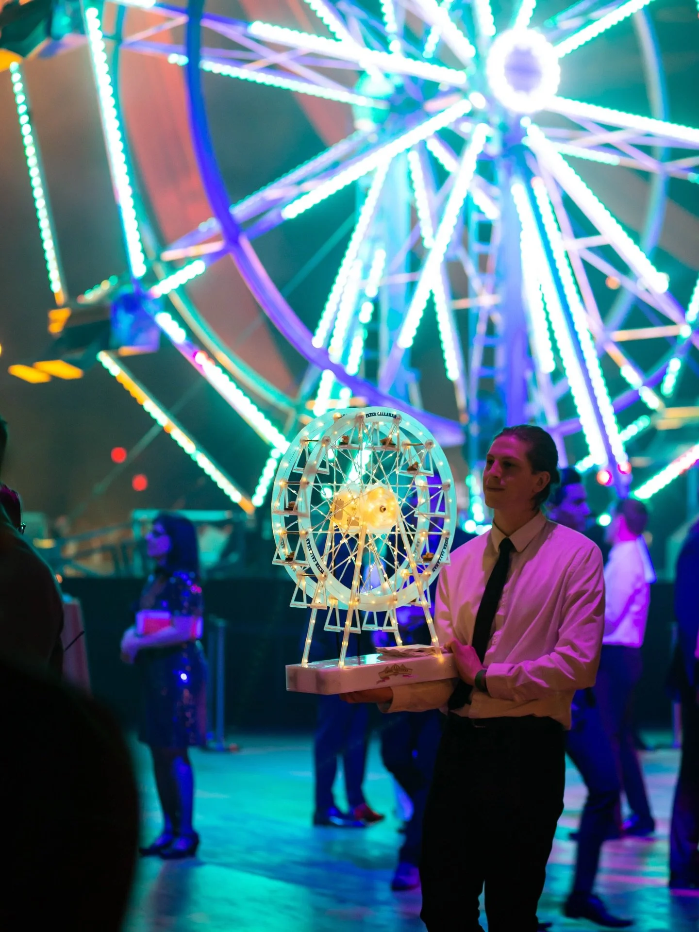 PCK server carrying a miniature Ferris wheel food display at a brightly lit evening event.