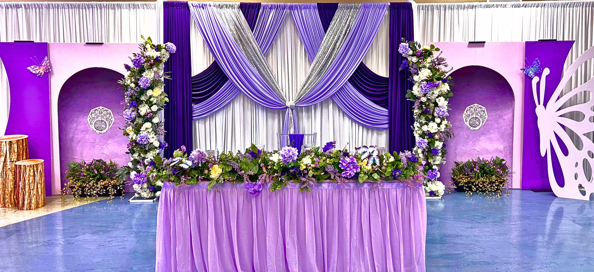 An Enchanted Sweet 16 brought to life with tree centerpieces, layered purple tones, and silver accents.   A life‑sized butterfly and a blooming garden backdrop complete this dreamy, storybook inspired celebration.