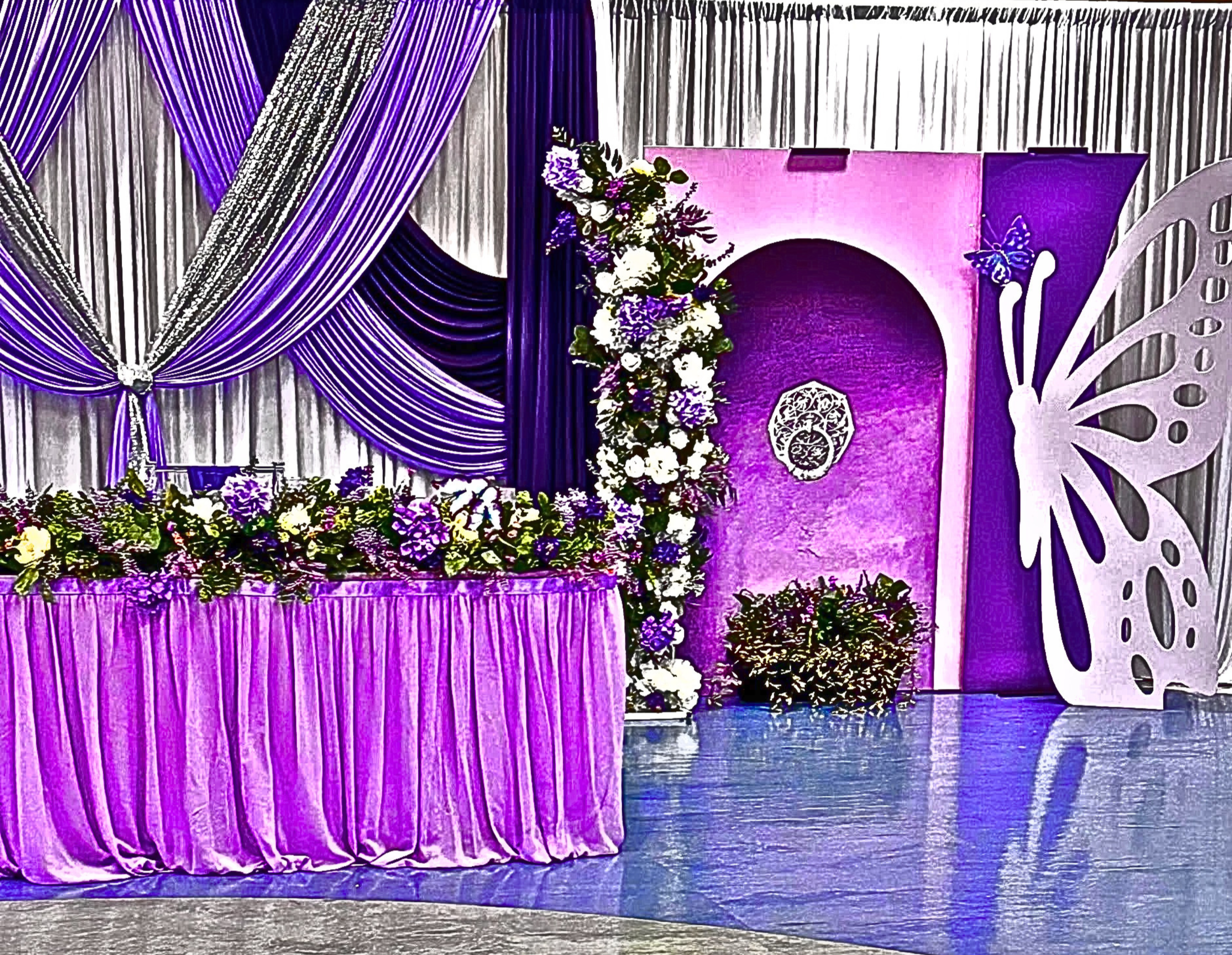 An Enchanted Sweet 16 brought to life with tree centerpieces, layered purple tones, and silver accents.   A life‑sized butterfly and a blooming garden backdrop complete this dreamy, storybook inspired celebration.