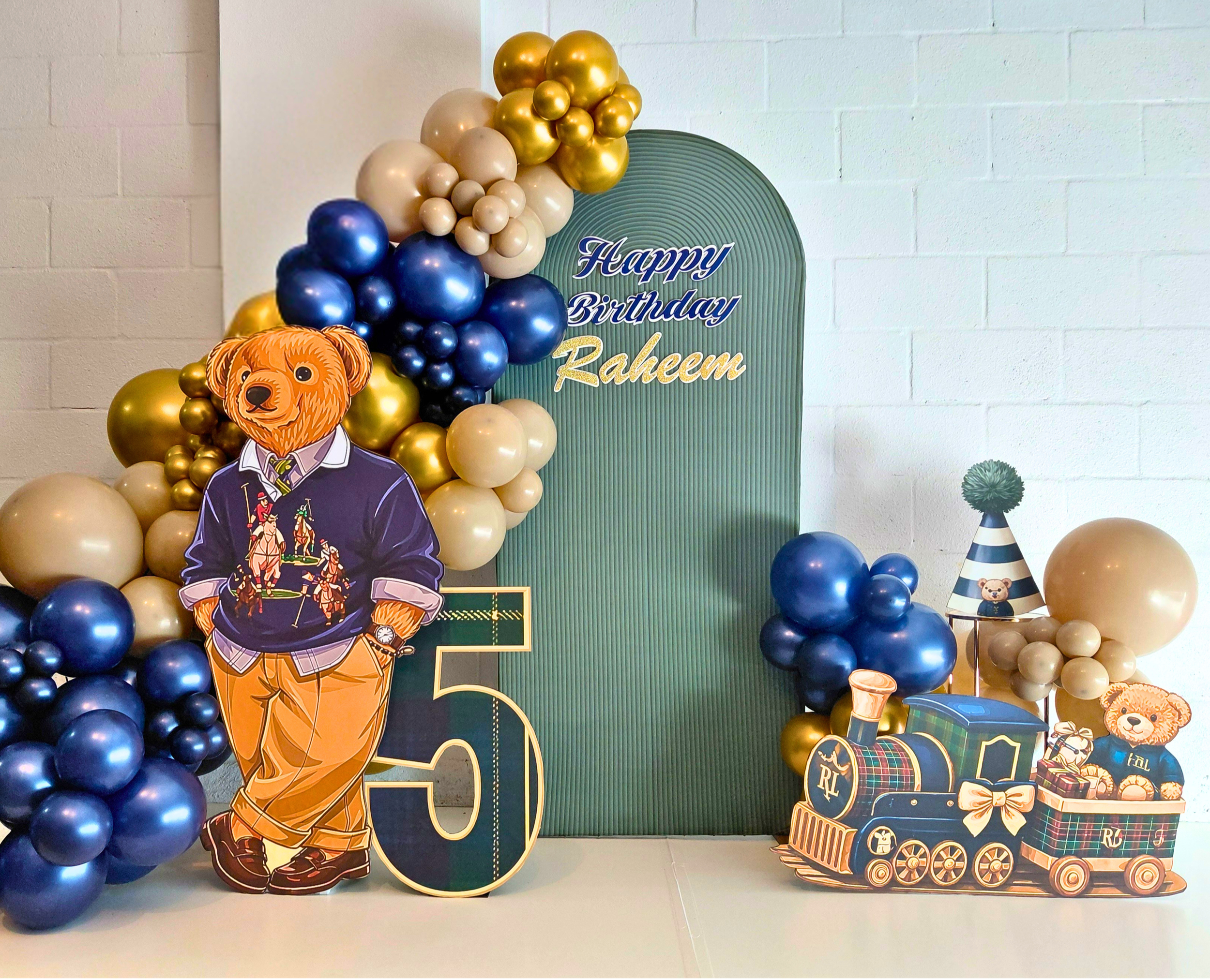 A charming Teddy Bear–themed 5th birthday celebration designed with bold blue, tan, and gold color‑blocking.  A life‑sized bear, mini train, birthday hat, and number 5 prop highlight the theme and complete the cohesive look.
