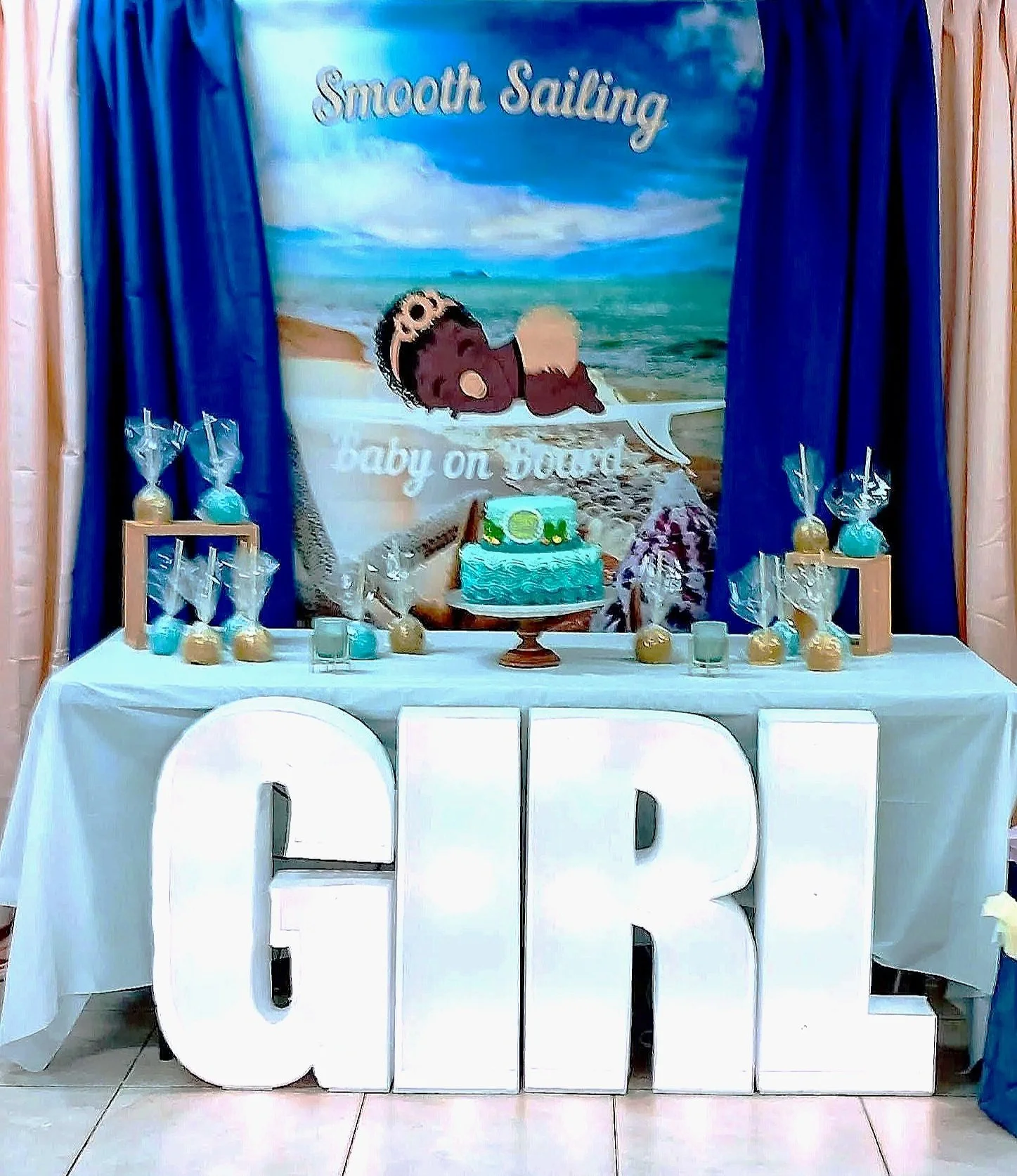 A baby shower setup with a beach-themed backdrop that says "Smooth Sailing". There is a cake and decorations on a table with a large 'BABY' in white letters at the front.