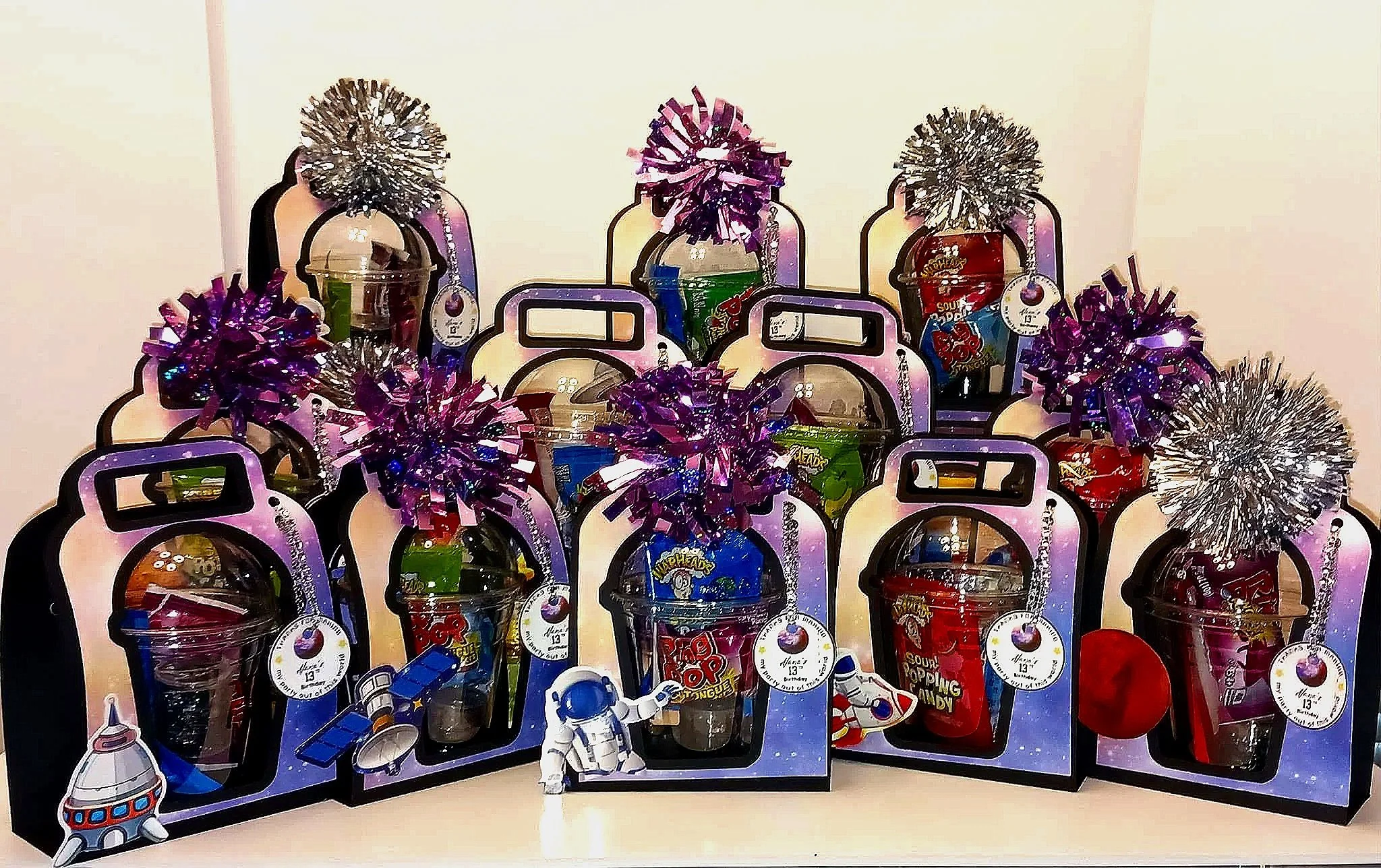 Collection of colorful toy bubble poppers shaped like bottles, decorated with metallic tinsel pom-poms in purple and silver, along with a small astronaut figurine at the bottom front.