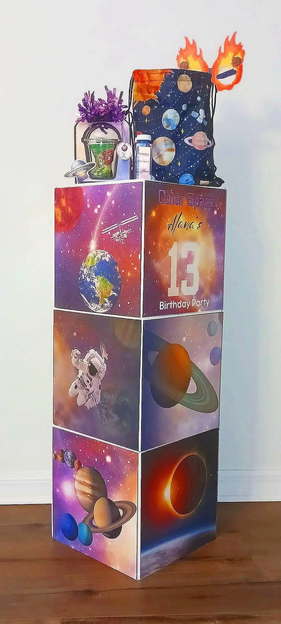 A birthday party decoration with a space theme, featuring building blocks with images of planets, galaxies, astronauts, and a spaceship, labeled "Outer Space," with a purple bucket of purple flowers on top.