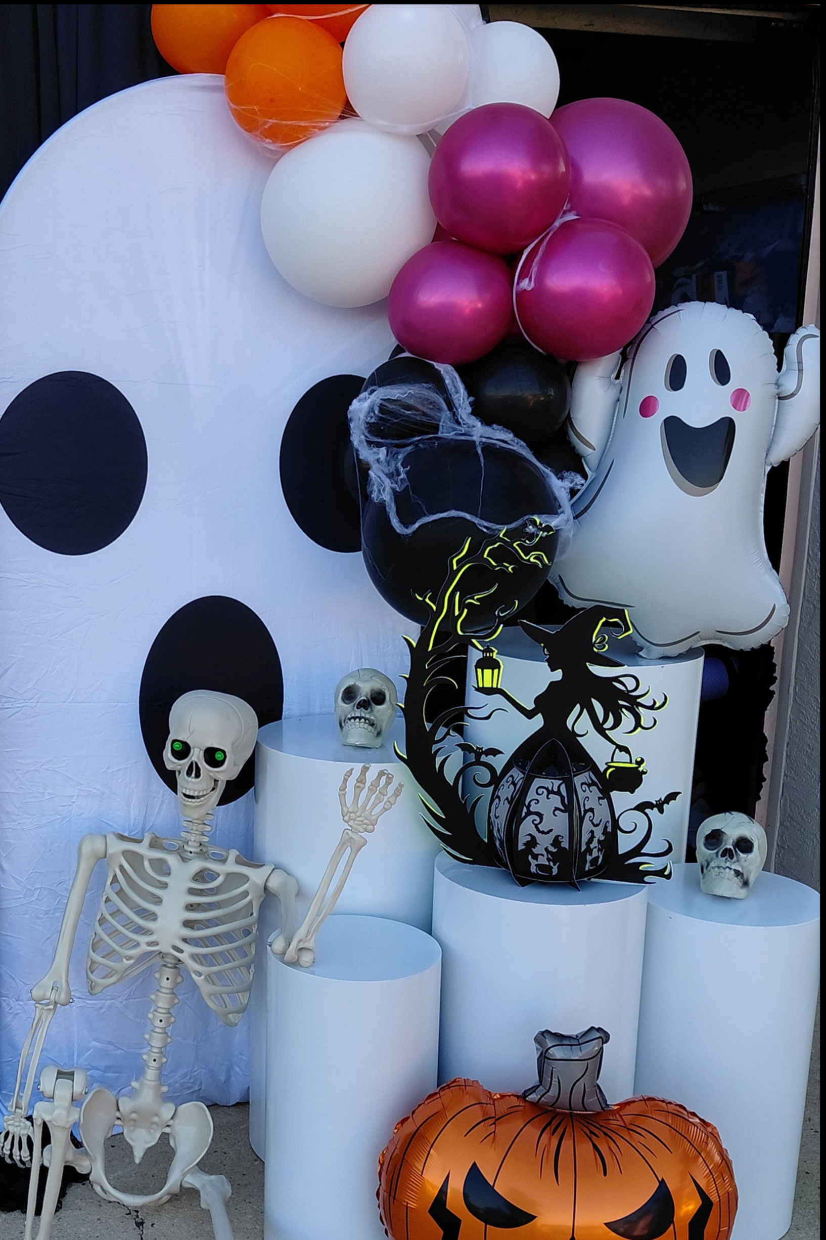 Halloween decorations featuring a large black cat poster with a pole, a white skeleton figure on a white cylindrical stand, various skull figures, orange and purple balloons, a black and orange pumpkin balloon at the bottom, and a ghost-shaped balloo