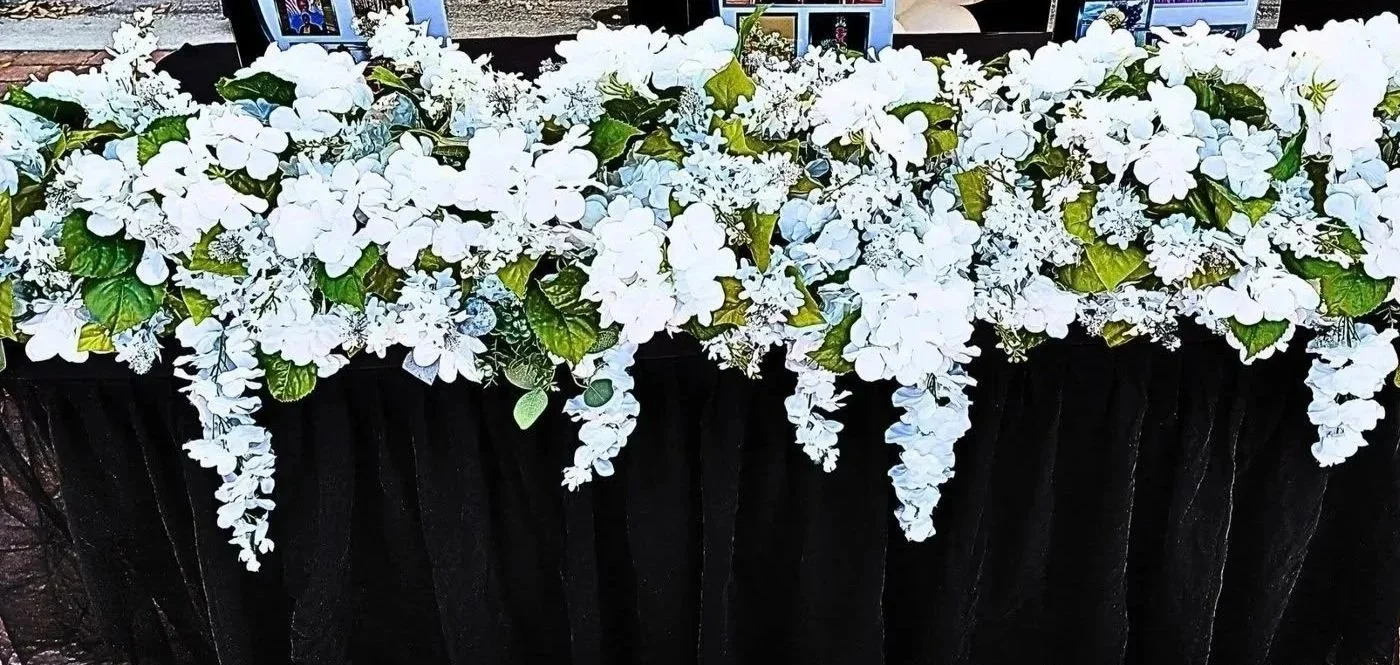 A display of white flowers with green leaves in a rectangular container wrapped with black material.