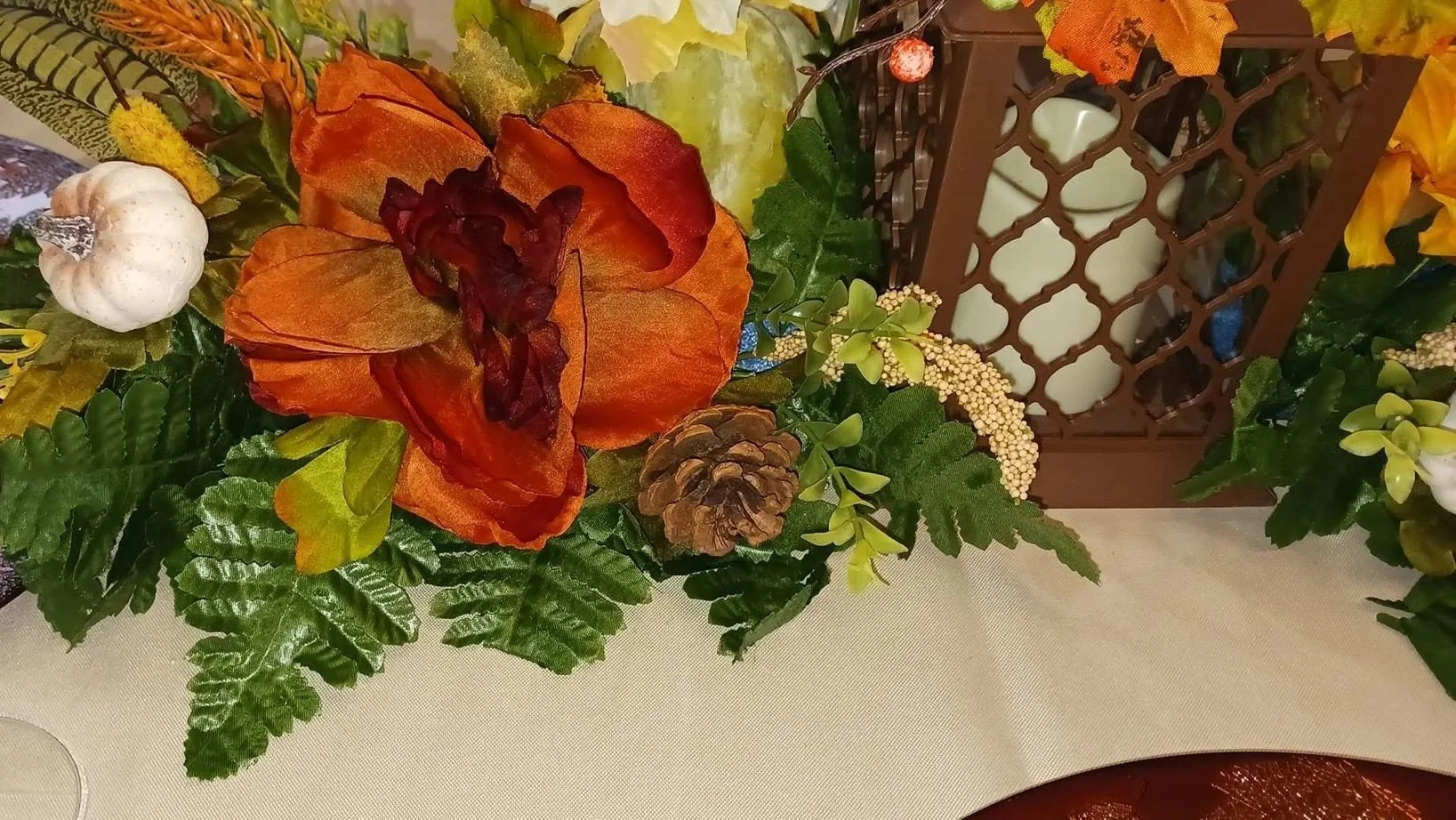 Autumn-themed floral arrangement with orange and red flowers, green leaves, a small white pumpkin, neighboring white pumpkin, pine cone, and yellow berries, with a brown lattice candle holder in the background, resting on a cream-colored tablecloth.