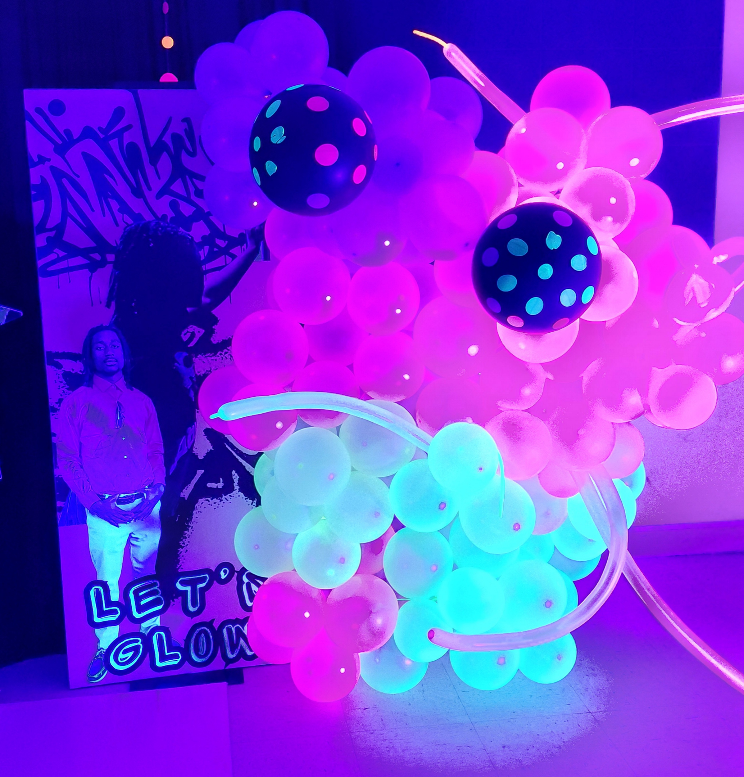 Colorful glowing balloons in pink, purple, and blue hues, with a sign that says "Let's Glow" and a silhouette of a person taking a photo, creating a vibrant, neon-lit scene.