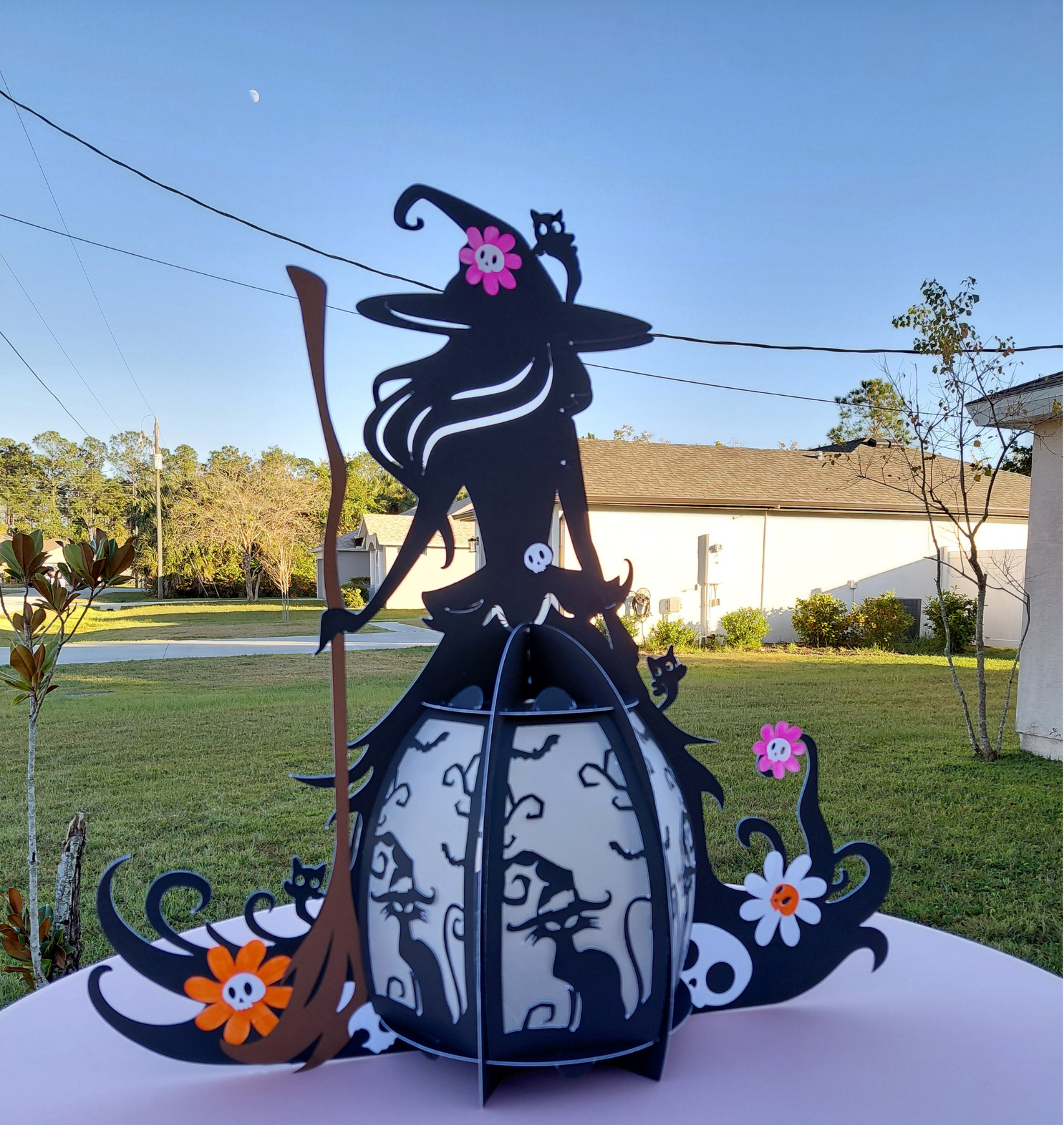 Paper Halloween decoration featuring a black cat with a pink flower on its head, a black witch hat, and small orange and white flowers, set against an outdoor background with a house, trees, and a clear blue sky.