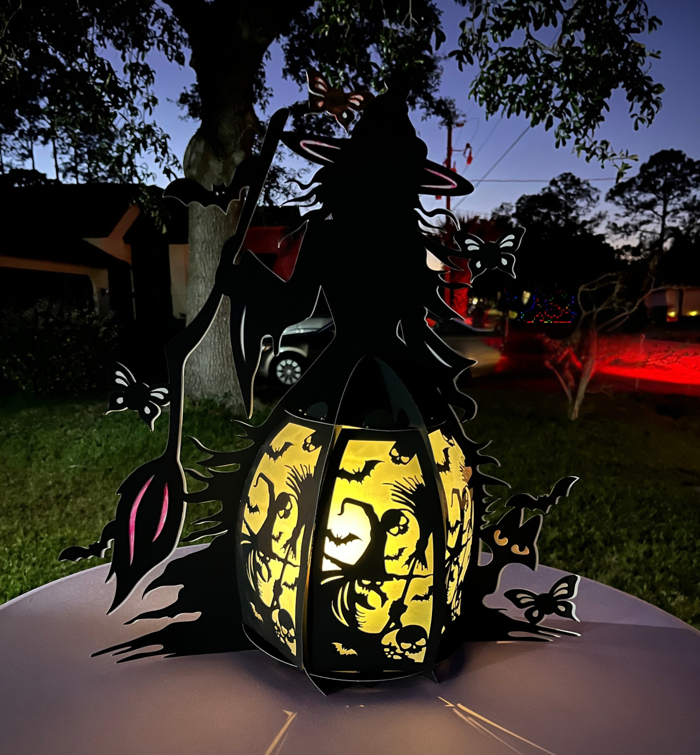 Silhouette of a decorative spooky pumpkin lantern with carved face, illuminated from within, surrounded by metal cutouts of bats and witches, set outdoors during twilight with trees and power lines in the background.