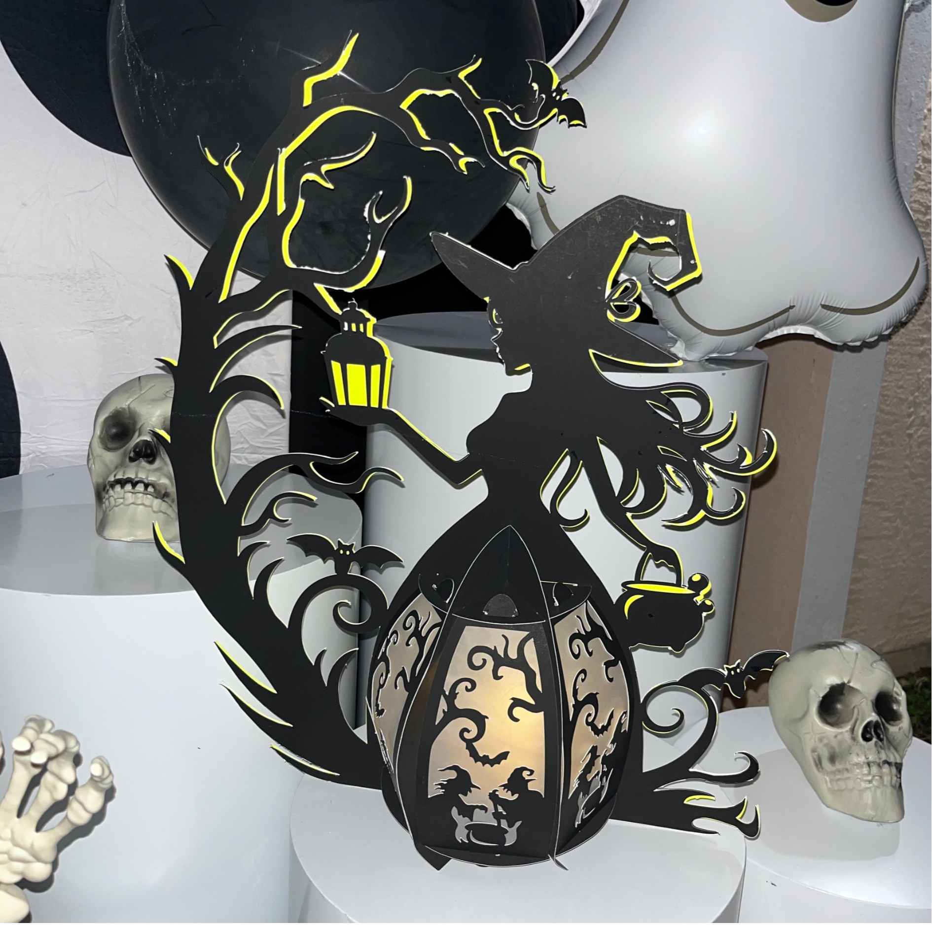 Decorative Halloween-themed cutout featuring black and yellow silhouette designs of a tree, a haunted house, bats, a witch, and a moon, accompanied by skeleton decorations and white balloons.
