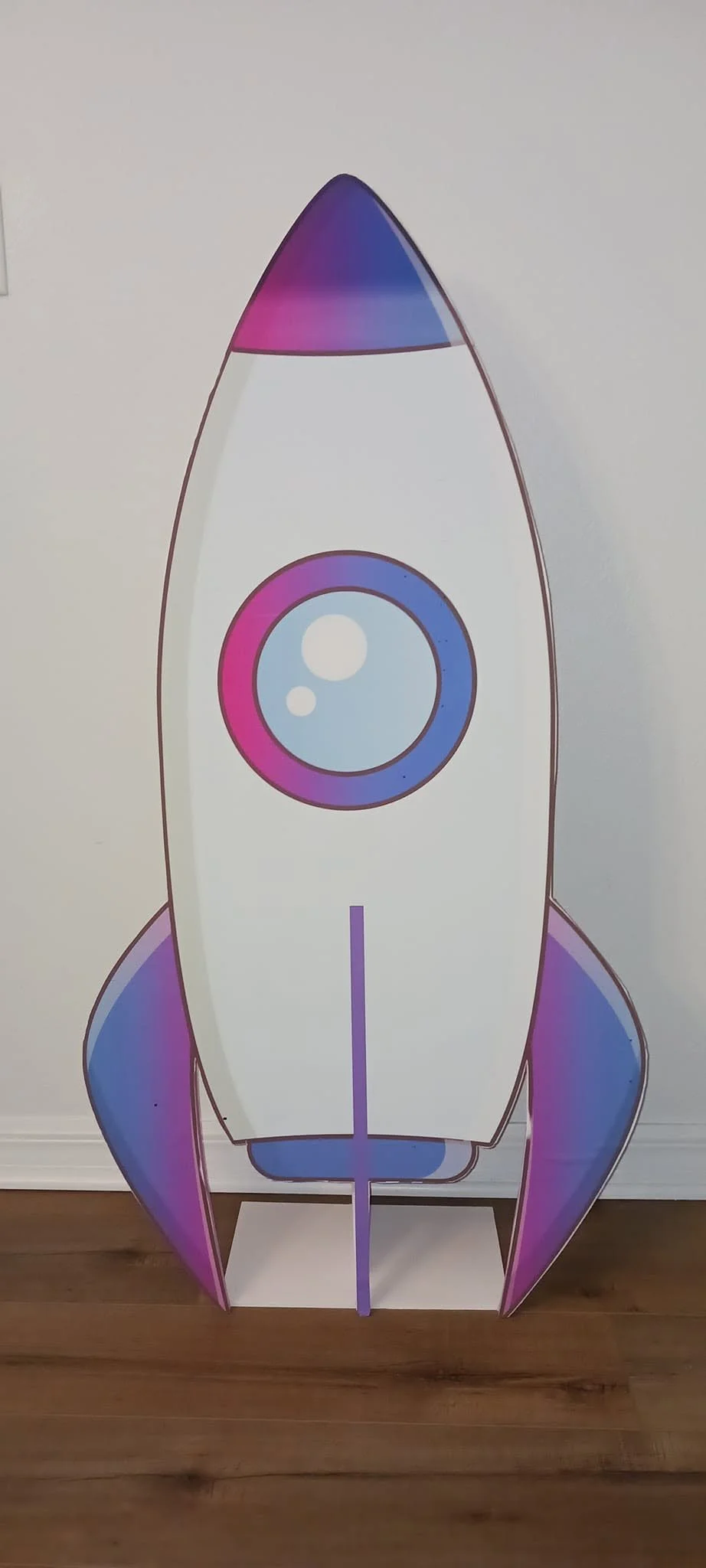 Colorful cardboard rocket ship decoration with purple, pink, and blue accents, standing on a wooden floor in front of a white wall.
