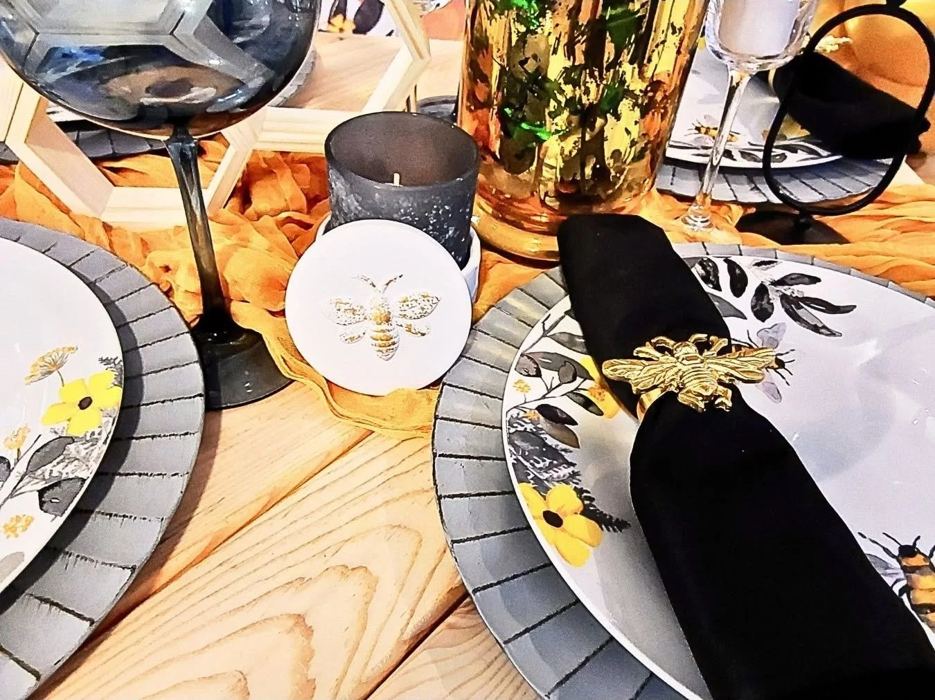 Table setting with bee-themed decorations, including plates with bee and flower designs, black napkin with a gold bee napkin ring, wine glasses, candles, and a large amber-colored vase with greenery, on a wooden table with an orange table runner.