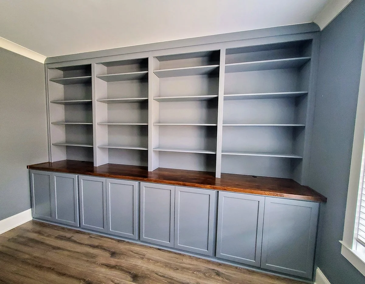 Empty built-in bookshelf with light gray shelves and cabinets with a dark wood countertop in a room with gray walls and wood flooring.