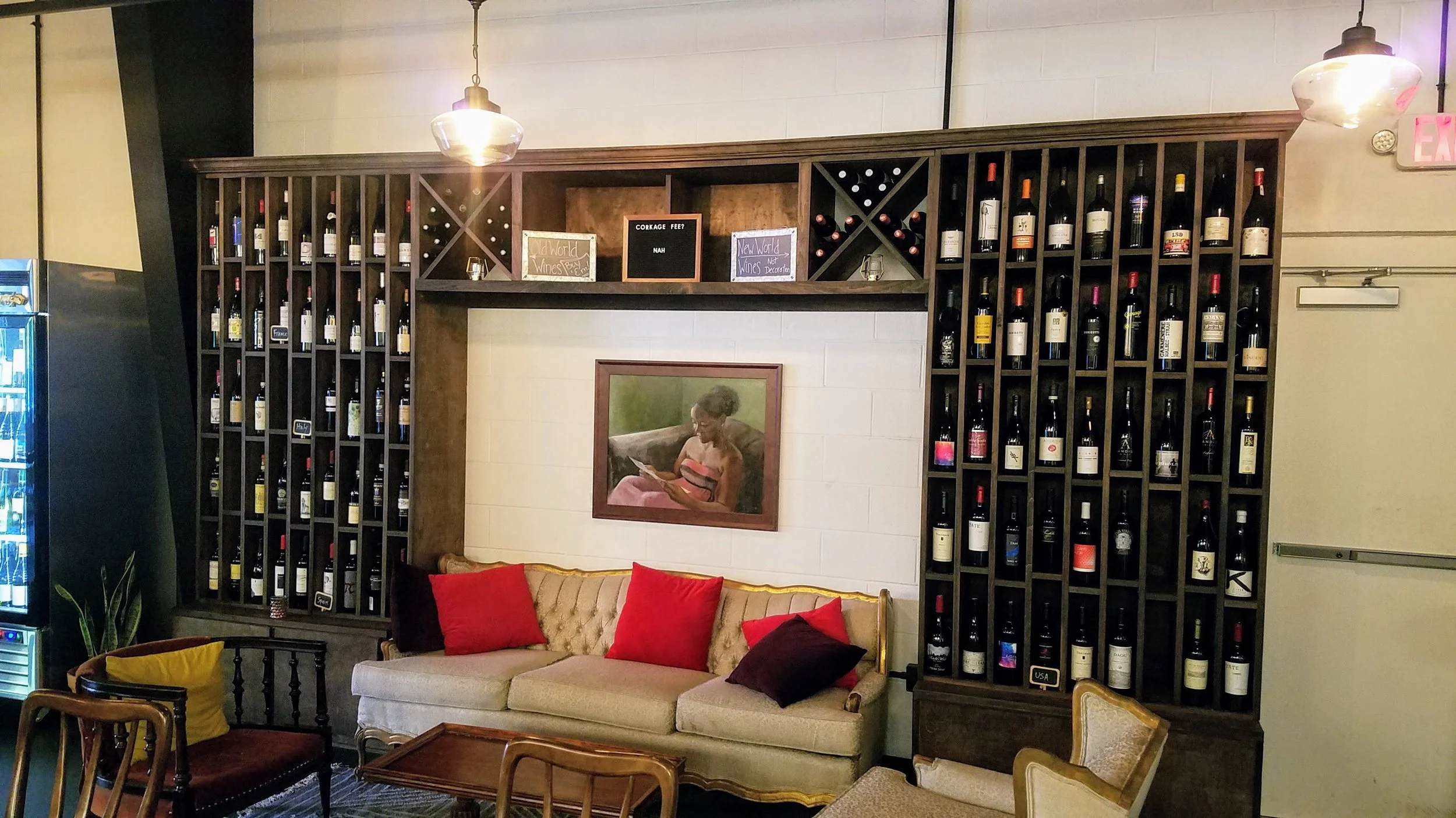 A cozy living room with a beige sofa adorned with red and dark pillows, facing a coffee table, against a white wall. Above, a large wooden wine rack filled with wine bottles spans the wall. A framed painting of a woman reading is centered on the wall below the rack. Hanging light fixtures illuminate the space, and part of a vending machine is visible on the left, with an exit sign on the right.