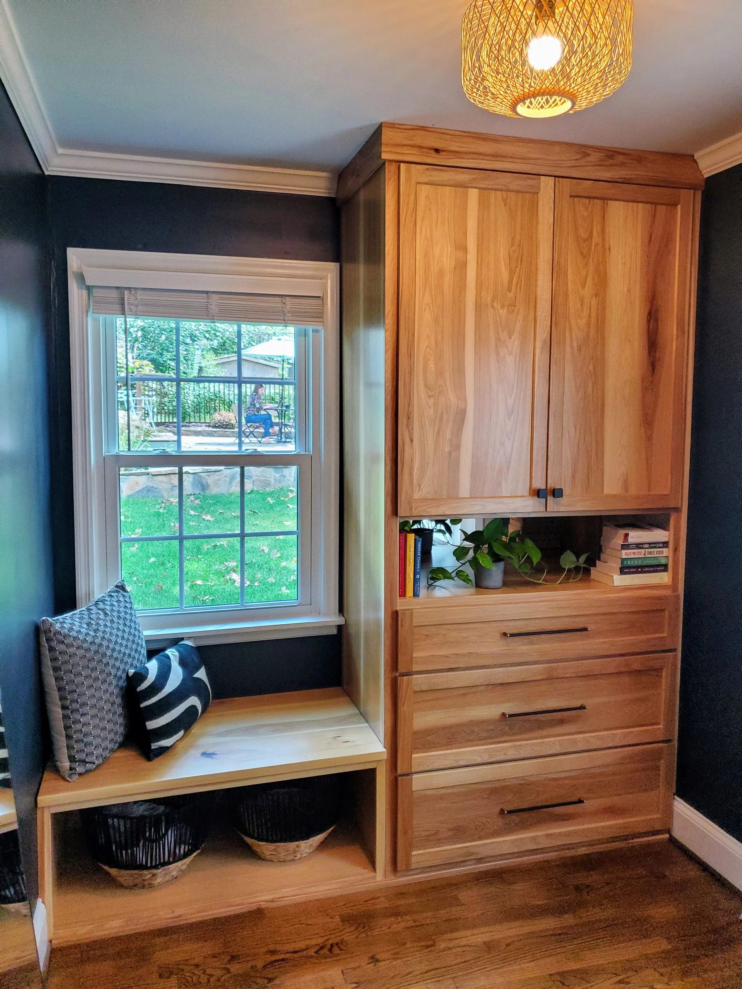 Interior view of a room with a window showing a backyard with a woman sitting at a table outside. Inside, a wooden cabinet and a bench with pillows; baskets and books are on the shelves.