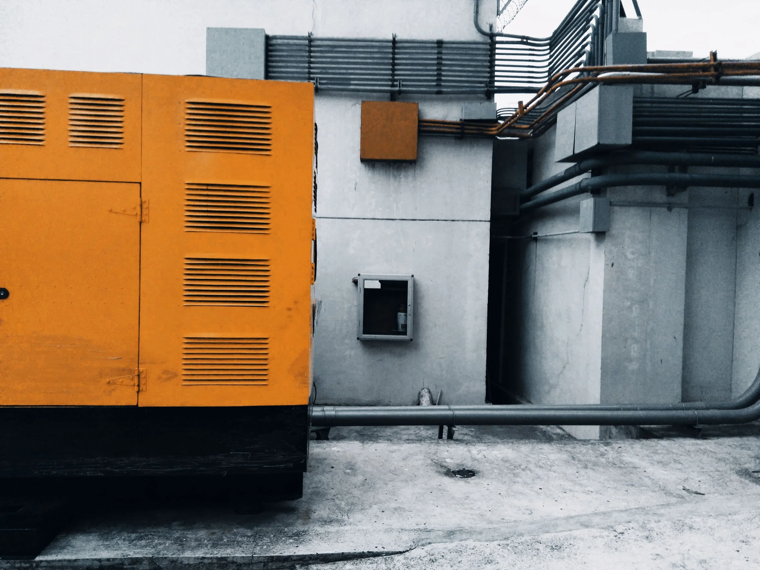 An exterior view of industrial equipment on a rooftop, with a large orange unit and various pipes and vents against a gray concrete wall.