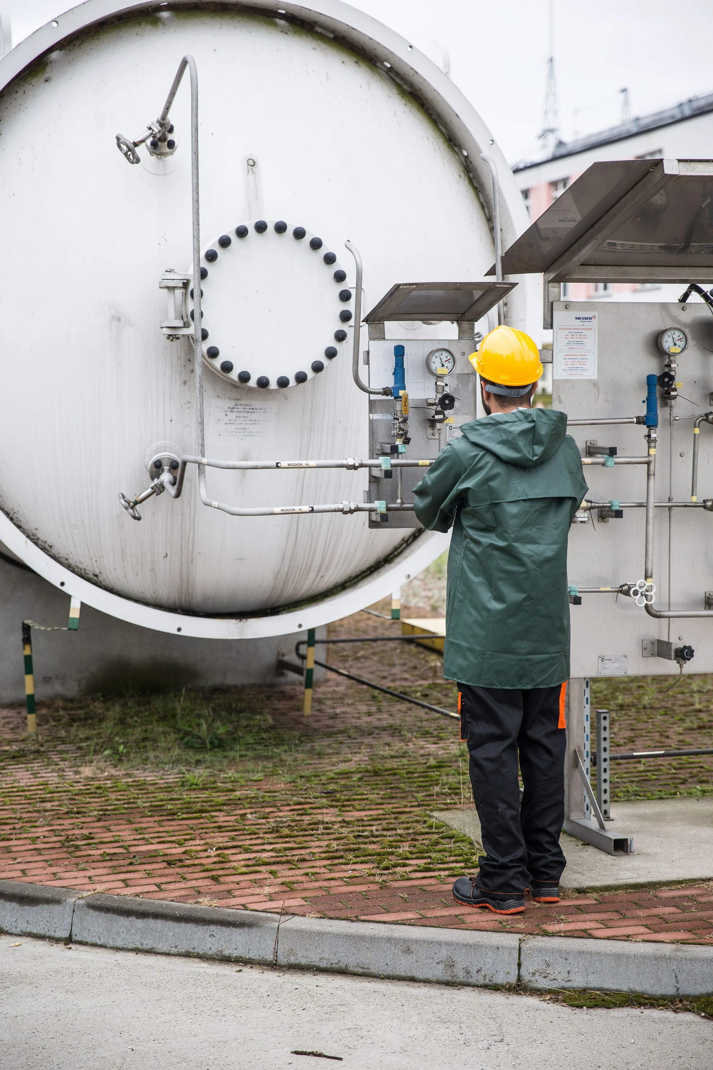 A worker wearing a yellow safety helmet and green jacket standing in front of a large, horizontal industrial tank with pipes, valves, and gauges. The tank is outdoors on a paved surface with grass and a building in the background.